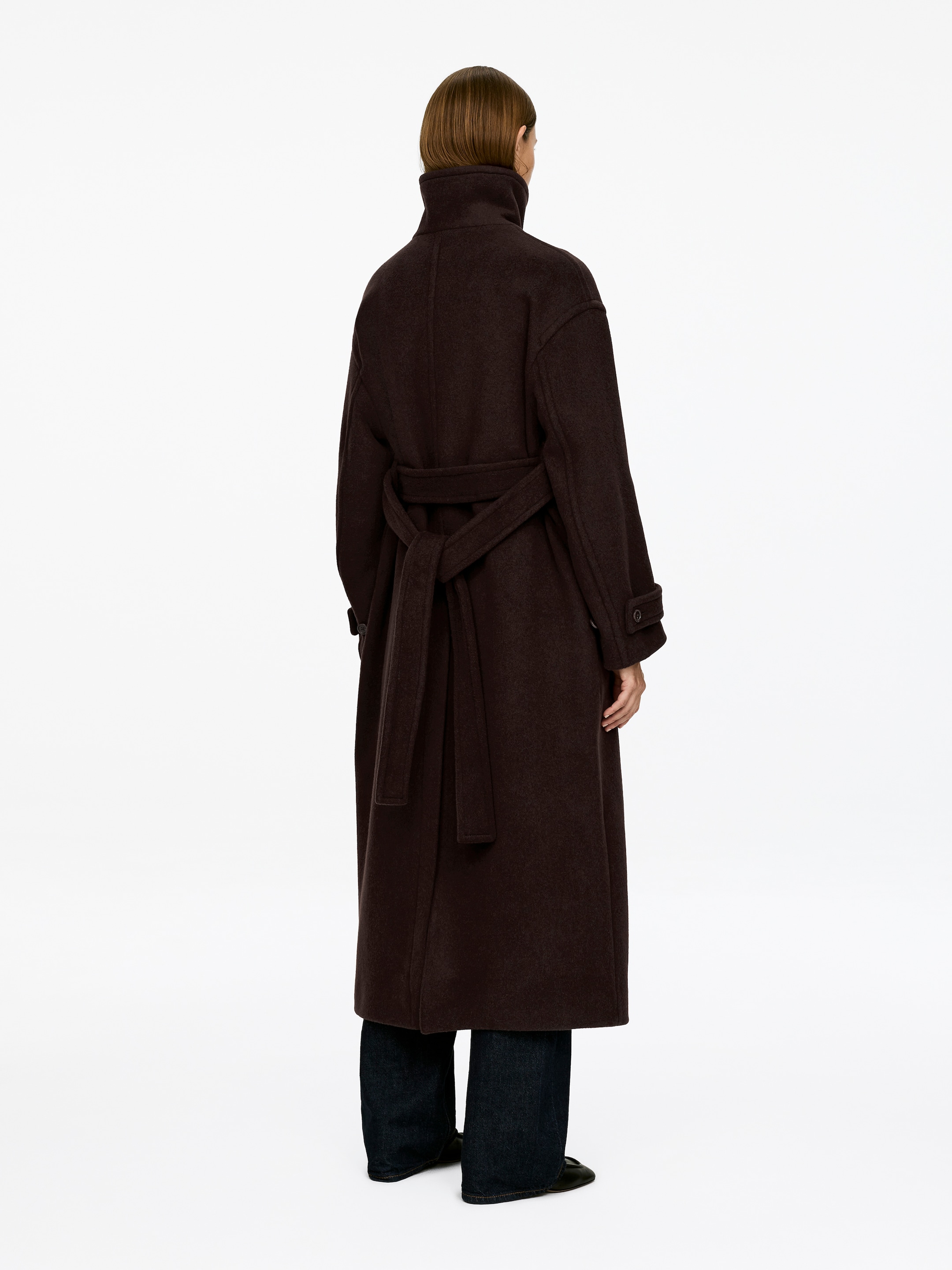 View larger image: A dark brown oversized belted maxi coat is being worn by the model, seen from the back. The coat features a standing collar and sleeve tabs with button closures, and its belt is tied at the back.