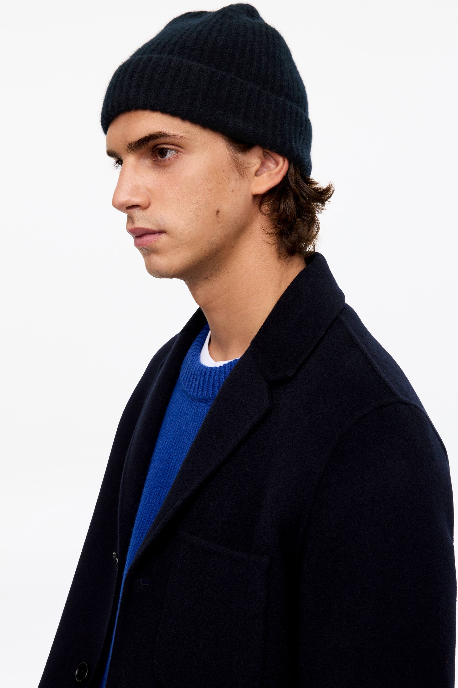 Visa större bild: En mörk marinblå stickad beanie med ribbad textur och uppvikt kant bärs av modellen. Beaniemössan stylas med en mörk marinblå kappa som bärs ovanpå en klarblå rundhalsad tröja.
