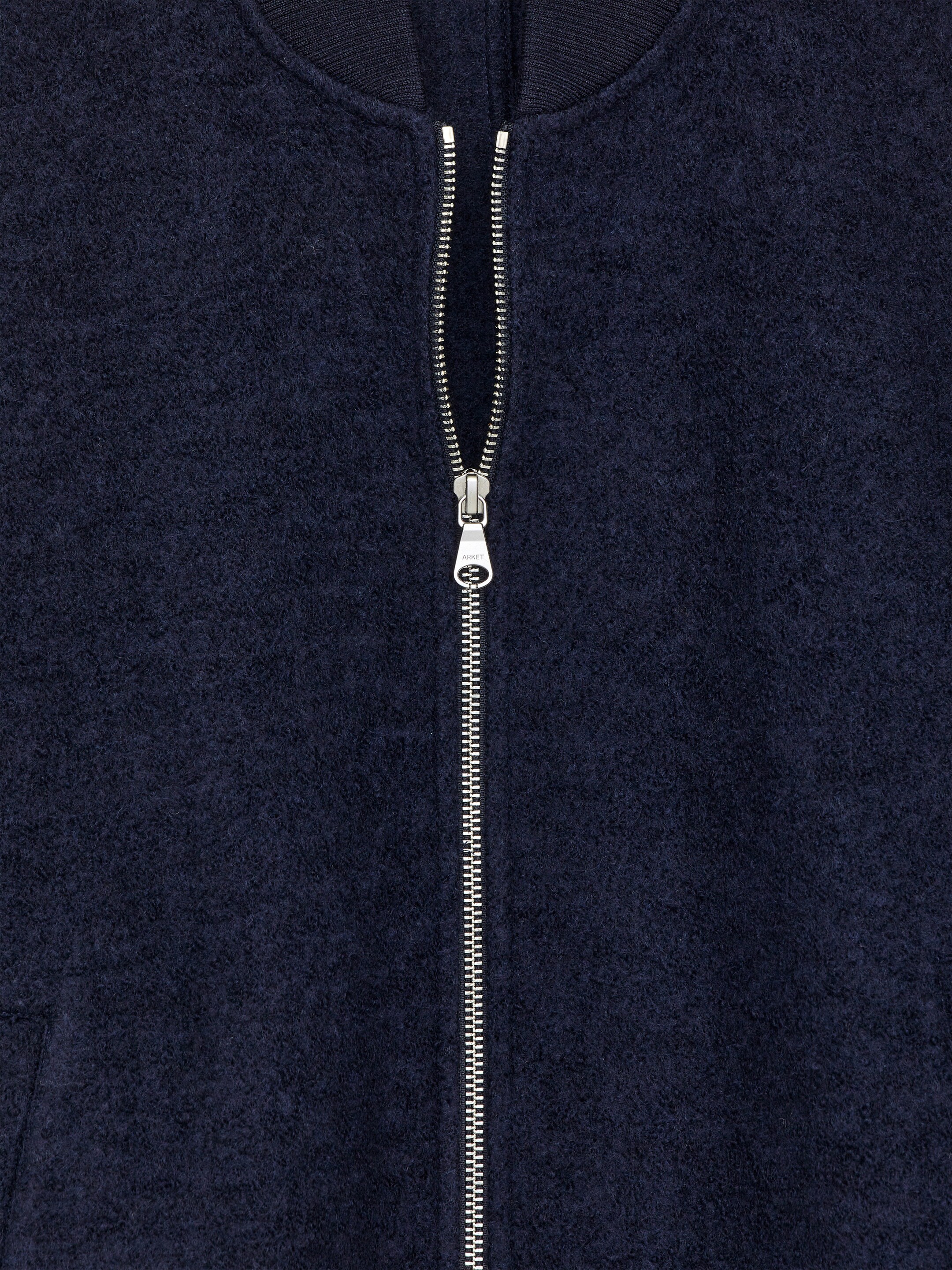 Größeres Bild anzeigen: Eine dunkelblaue Jacke, die von vorne gezeigt wird, zeigt einen auffälligen silbernen Zwei-Wege-Reißverschluss mit einem 'ARKET'-gebrandeten Zipper, der teilweise geöffnet ist. Die Jacke ist aus einer strukturierten Woll-Baumwoll-Mischung gefertigt, mit einem gerippten Kragen und einem Teil einer Leistentasche, die ebenfalls sichtbar ist.