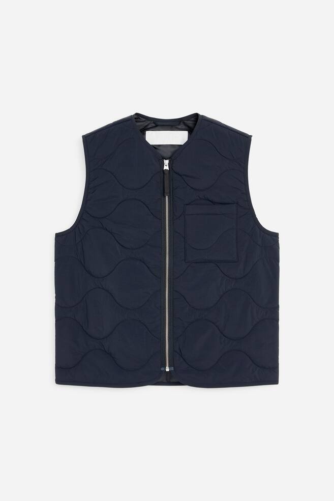 En mørkeblå polstret vest præsenteres forfra og har et gennemgående mønster af løgformet quiltning. Den er designet med en sølvfarvet lynlås foran og en påsyet lomme på bærerens venstre side. - 1