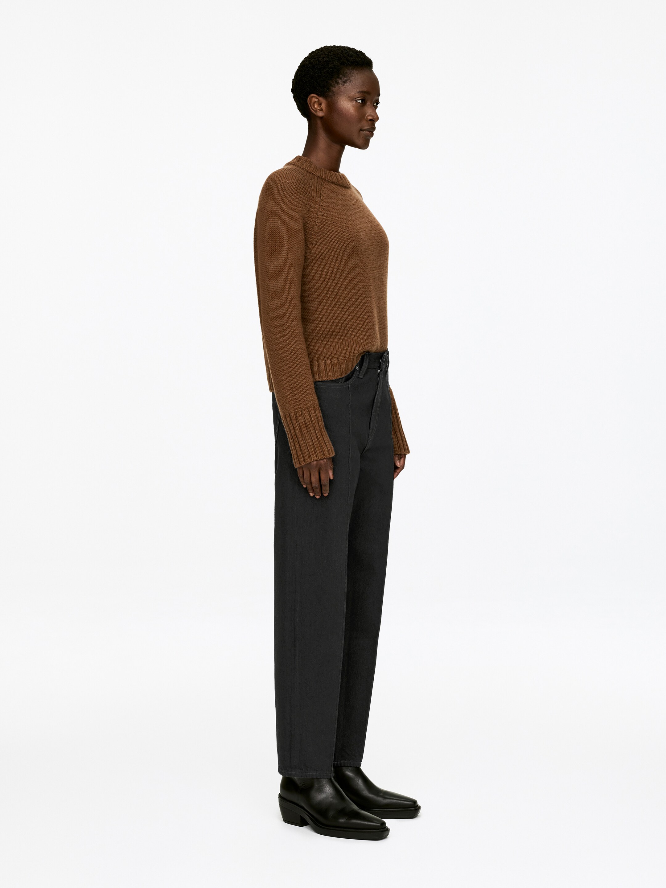 Agrandir l'image: Un pull à col rond en maille marron présente des manches raglan, des poignets côtelés et un ourlet côtelé, porté de profil, tourné vers la droite. Il est associé à un pantalon droit gris foncé et à des bottines noires à bout pointu.