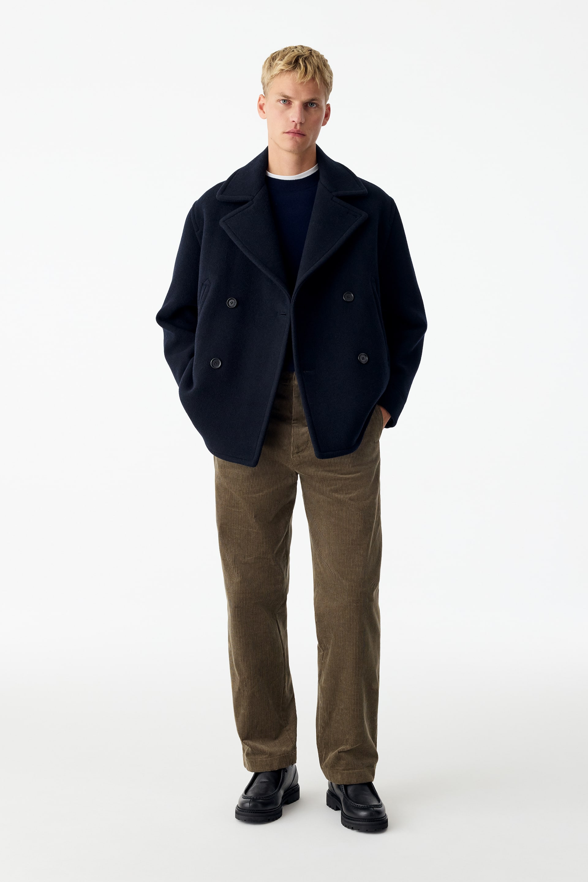 View larger image: Brown corduroy trousers in a relaxed, wide-leg fit are being worn by the model. The trousers are styled with a navy double-breasted jacket, a dark blue crew-neck sweater, and black leather shoes.