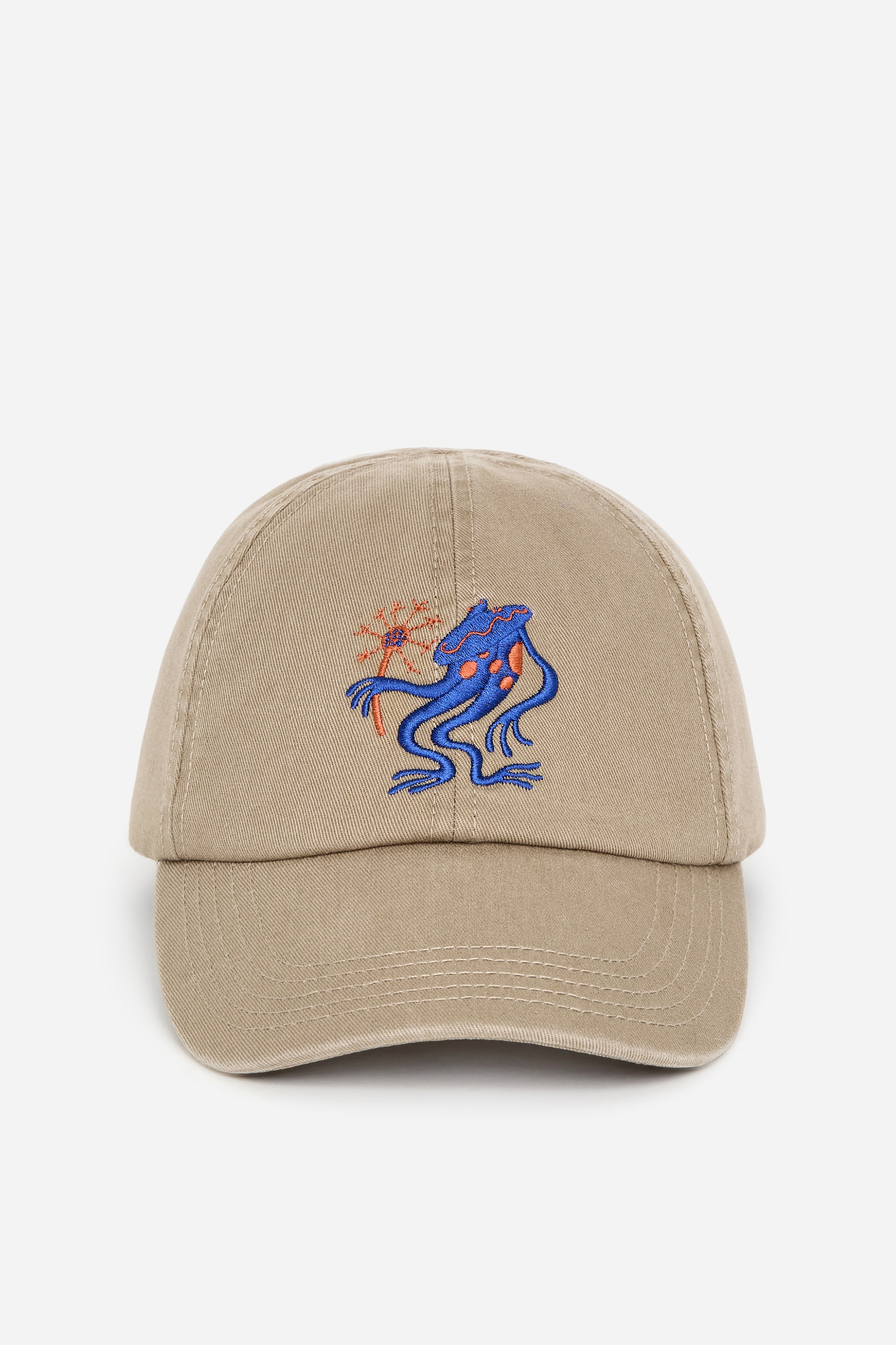 Ingrandisci l'immagine: Un berretto color camoscio in stile baseball è mostrato frontalmente, caratterizzato da dettagli ricamati. Sul pannello frontale è raffigurata una creatura stilizzata di colore blu con accenti arancioni, che tiene in mano un oggetto appuntito di colore rosso-arancione.