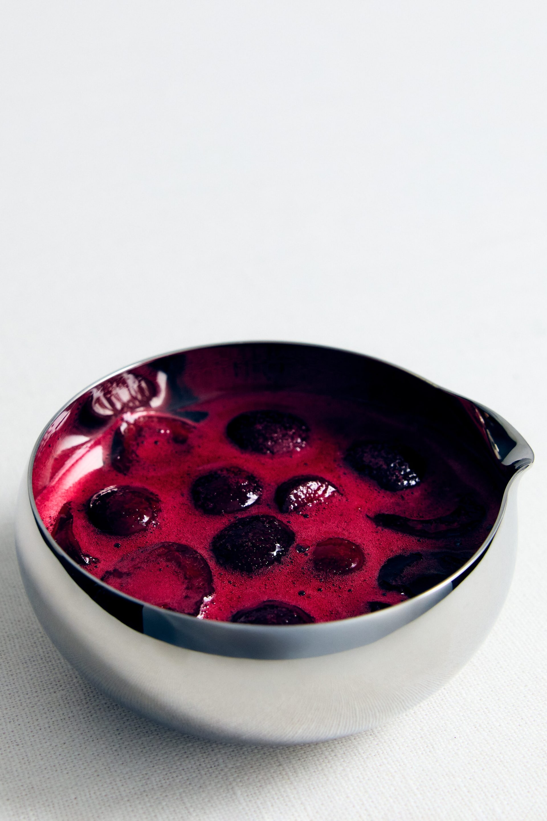 Visualizza immagine più grande: Una ciotola versatrice argentata e lucida contiene un vivace liquido rosso con frutti di bosco interi scuri che galleggiano in superficie, mostrando una leggera consistenza schiumosa.