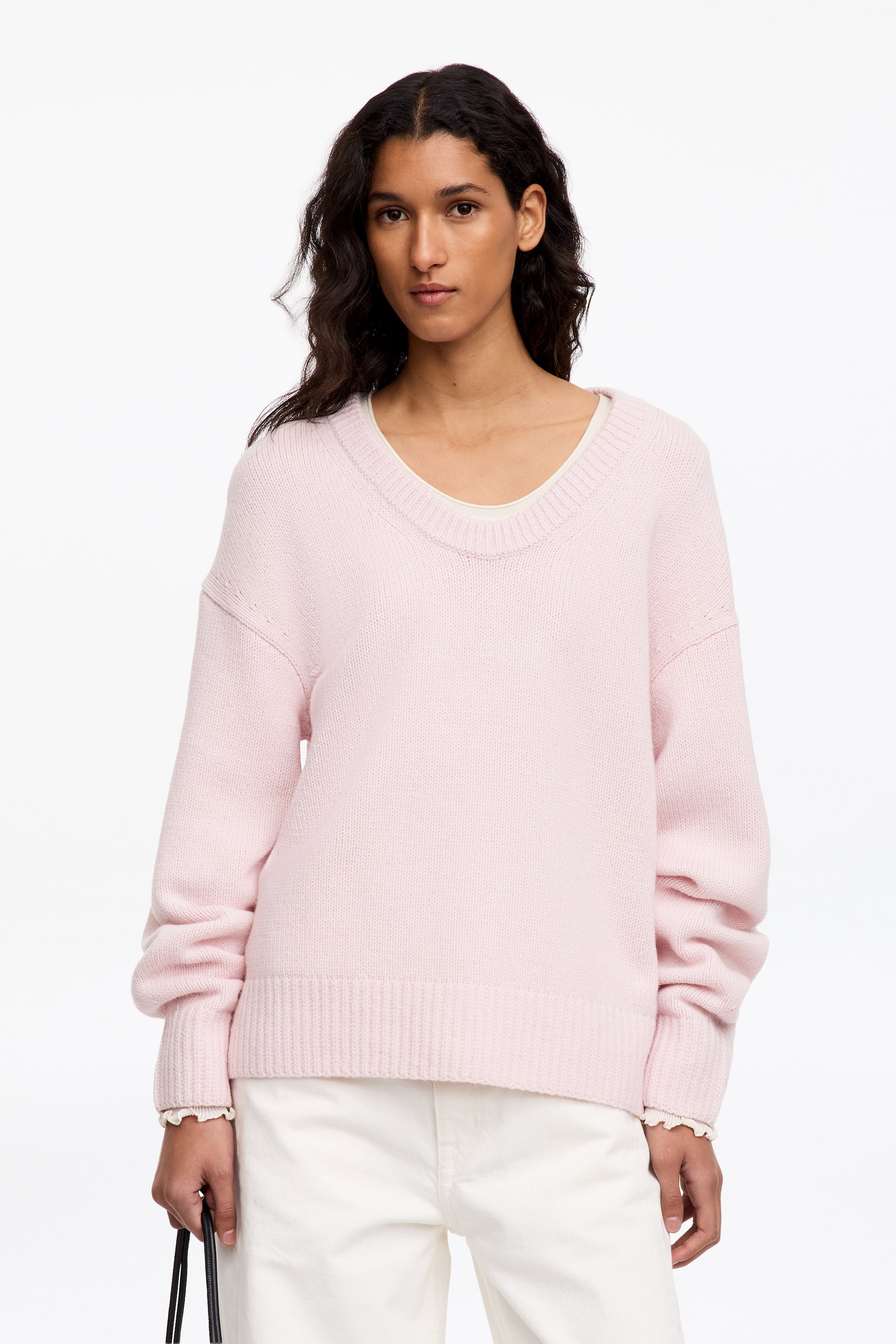 Agrandir l'image: Un pull en maille rose pâle avec un profond col en U, des bordures côtelées et des épaules tombantes est porté par le mannequin. Ce pull à la coupe ample est superposé sur un haut de couleur claire et associé à un pantalon blanc.