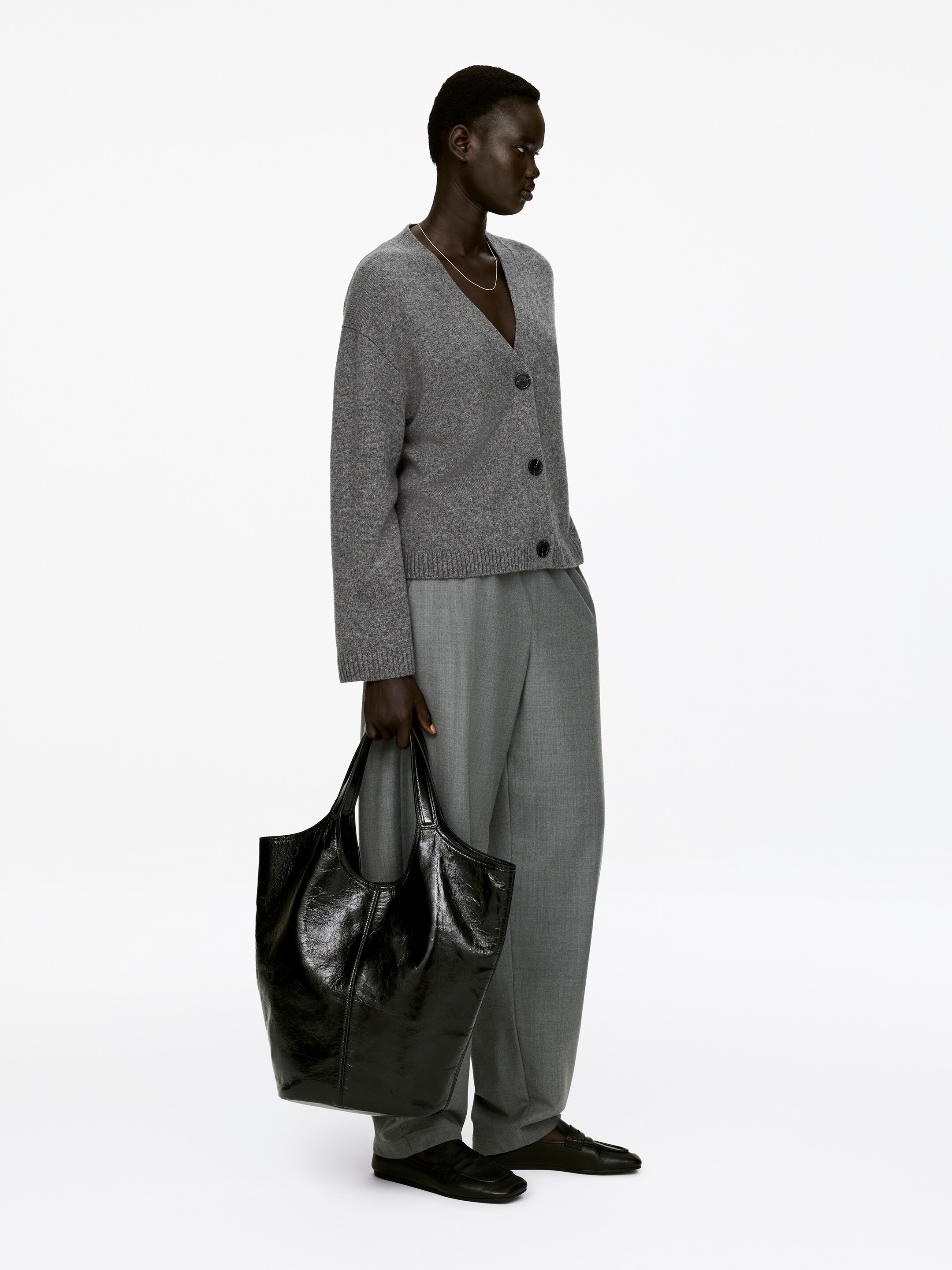 View larger image: A grey long-sleeved cardigan, featuring a V-neck, three visible buttons, and a rib-knitted hem, is being worn by the model. The cardigan is styled open, revealing a silver necklace, and is paired with grey trousers.