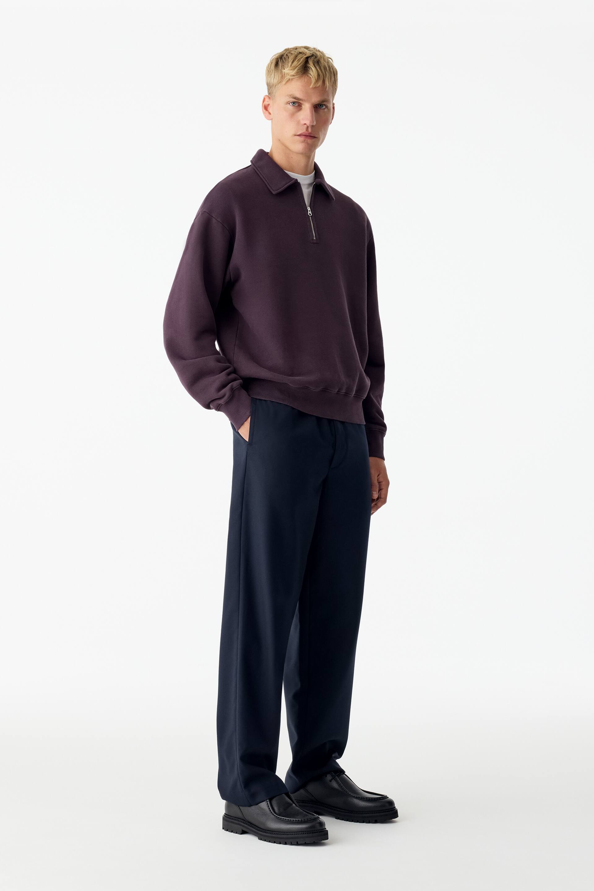 Grotere afbeelding bekijken: Een donkerpaarse sweatshirt, garment-dyed, met kraag, halve ritssluiting aan de voorkant en geribde afwerkingen, wordt gedragen door het model. De sweatshirt is gelaagd over een wit T-shirt en gestyled met een donkere broek.