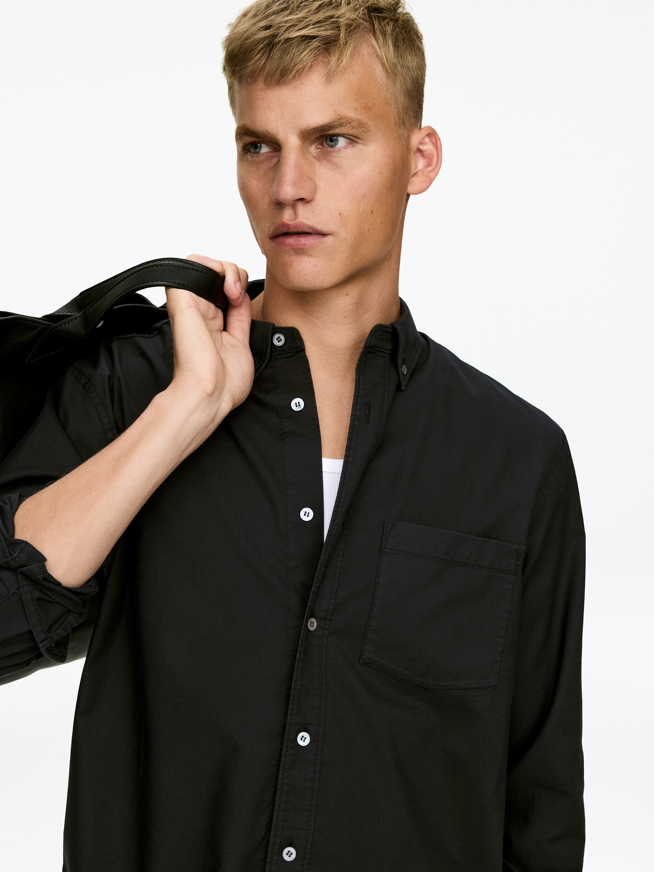 Ver imagen más grande: Una camisa negra de botones se está llevando puesta, inclinada ligeramente hacia la derecha, revelando un cuello de camisa de botones con botones blancos, una tapeta frontal con botones blancos y un bolsillo de parche en el pecho izquierdo. Sus mangas largas están enrolladas en el brazo visible, y una camiseta interior blanca se muestra debajo de la camisa parcialmente desabrochada.