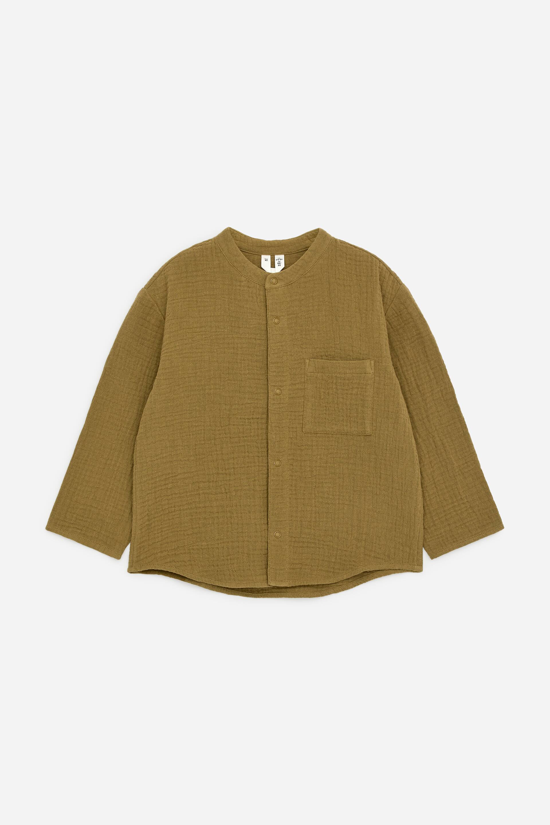 Преглед на по-голямо изображение: Риза без яка, представяща предната си част, изработена от памучен муселин в маслинено зелено, показваща текстурирана, намачкана повърхност. Характеризира се с яка тип "банд", закопчаване с копчета отпред, един джоб с капак на левия гръден кош и свободна кройка с дълги ръкави.