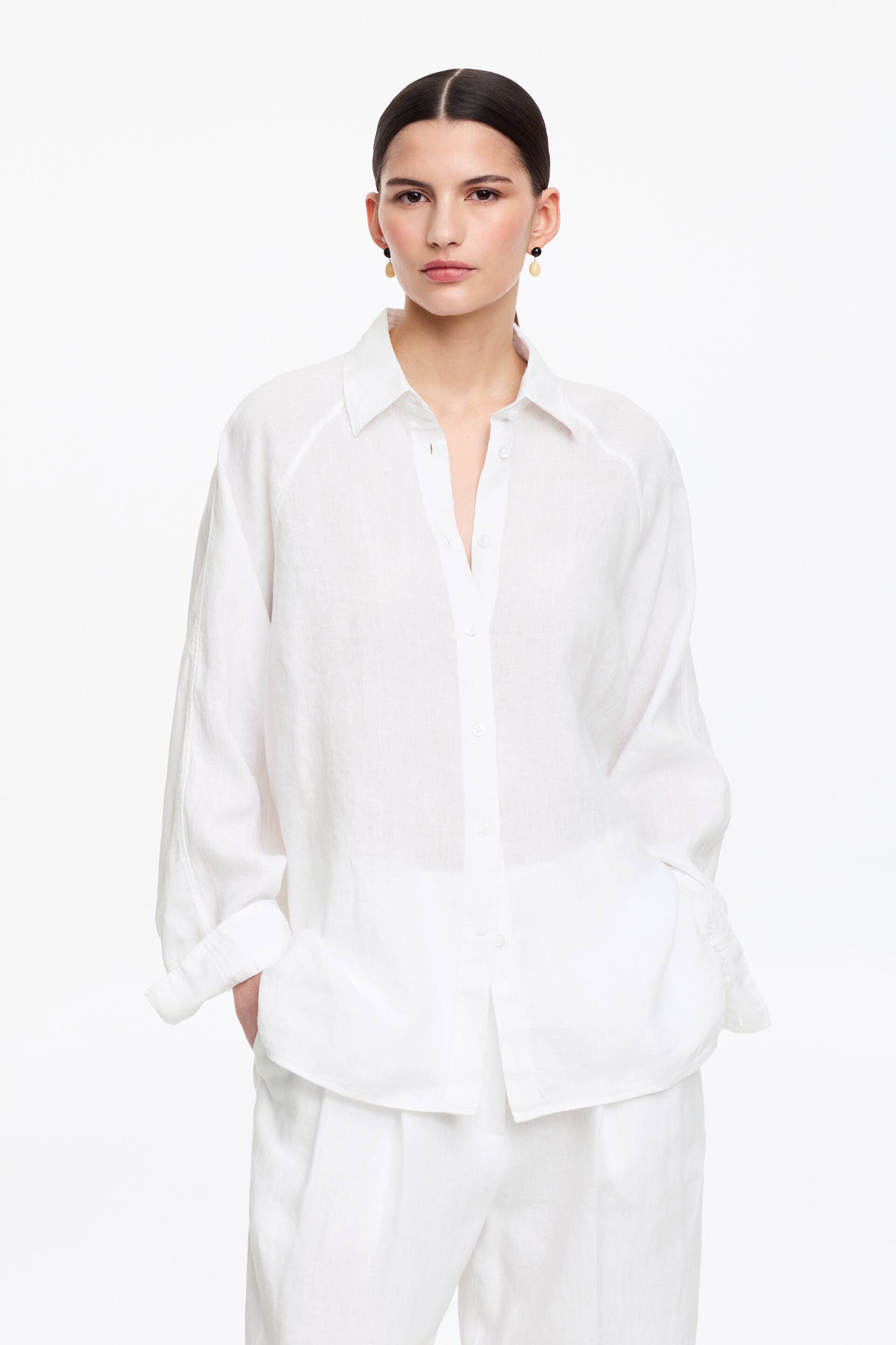 Näytä suurempi kuva: Valkoinen pellavainen nappipaita, jossa on terävä kaulus ja raglan-hihat, on puettu rennosti housujen päälle. Housut ovat valkoiset pellavaiset laskostetut housut. Paidan rento malli sisältää pitkät mansetilliset hihat, joista yksi on kääritty ylös.