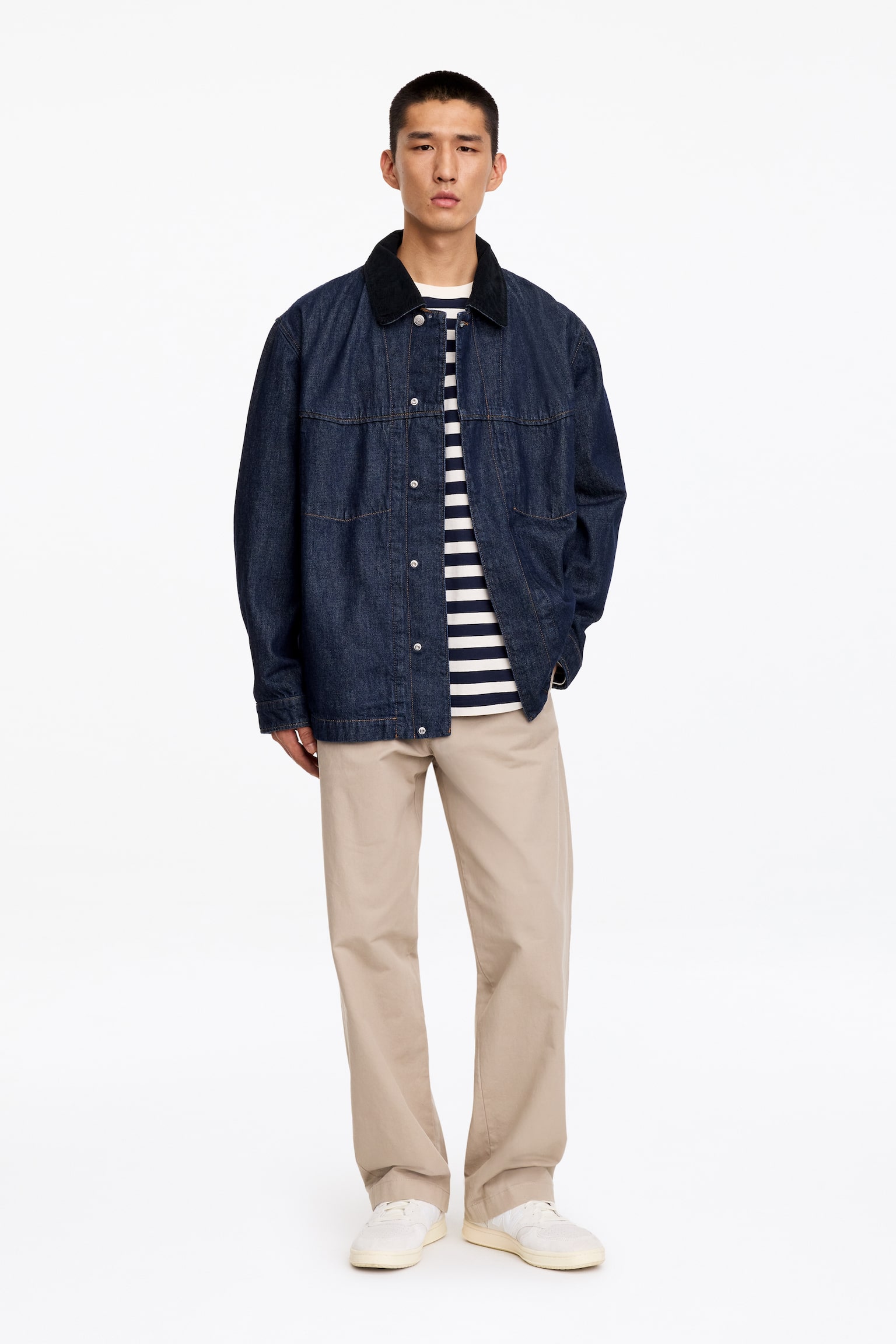 Une veste en denim de coupe décontractée, bleu foncé, dotée dâun col en velours côtelé noir et de coutures apparentes provenant des poches intérieures, est portée par le mannequin. La veste est stylée ouverte sur un t-shirt rayé bleu et blanc et un pantalon beige. - 1