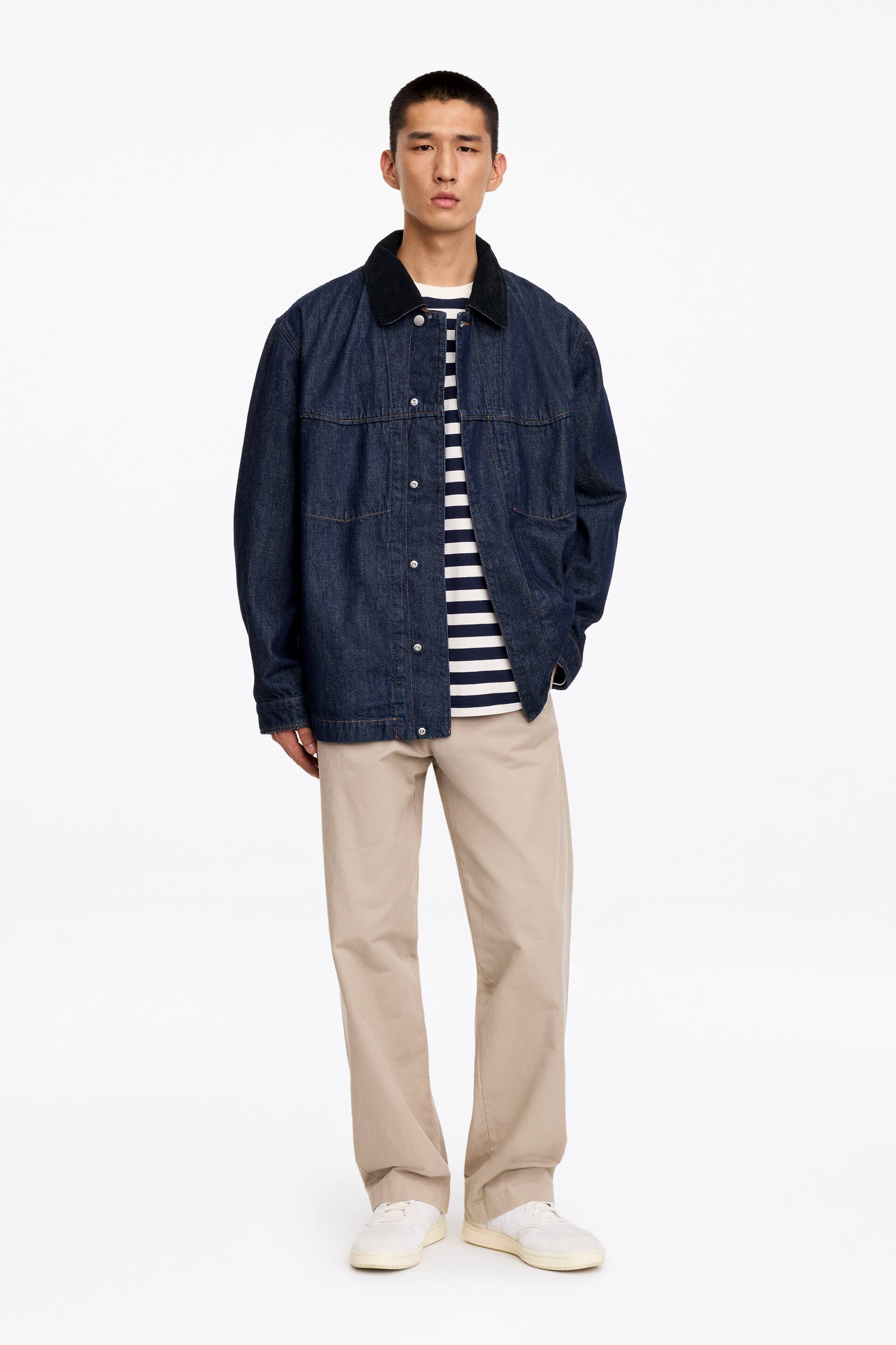 View larger image: A dark blue, relaxed-fit denim railway jacket featuring a black corduroy collar and visible stitching from internal pockets is being worn by the model. The jacket is styled open over a blue-and-white striped T-shirt and beige trousers.