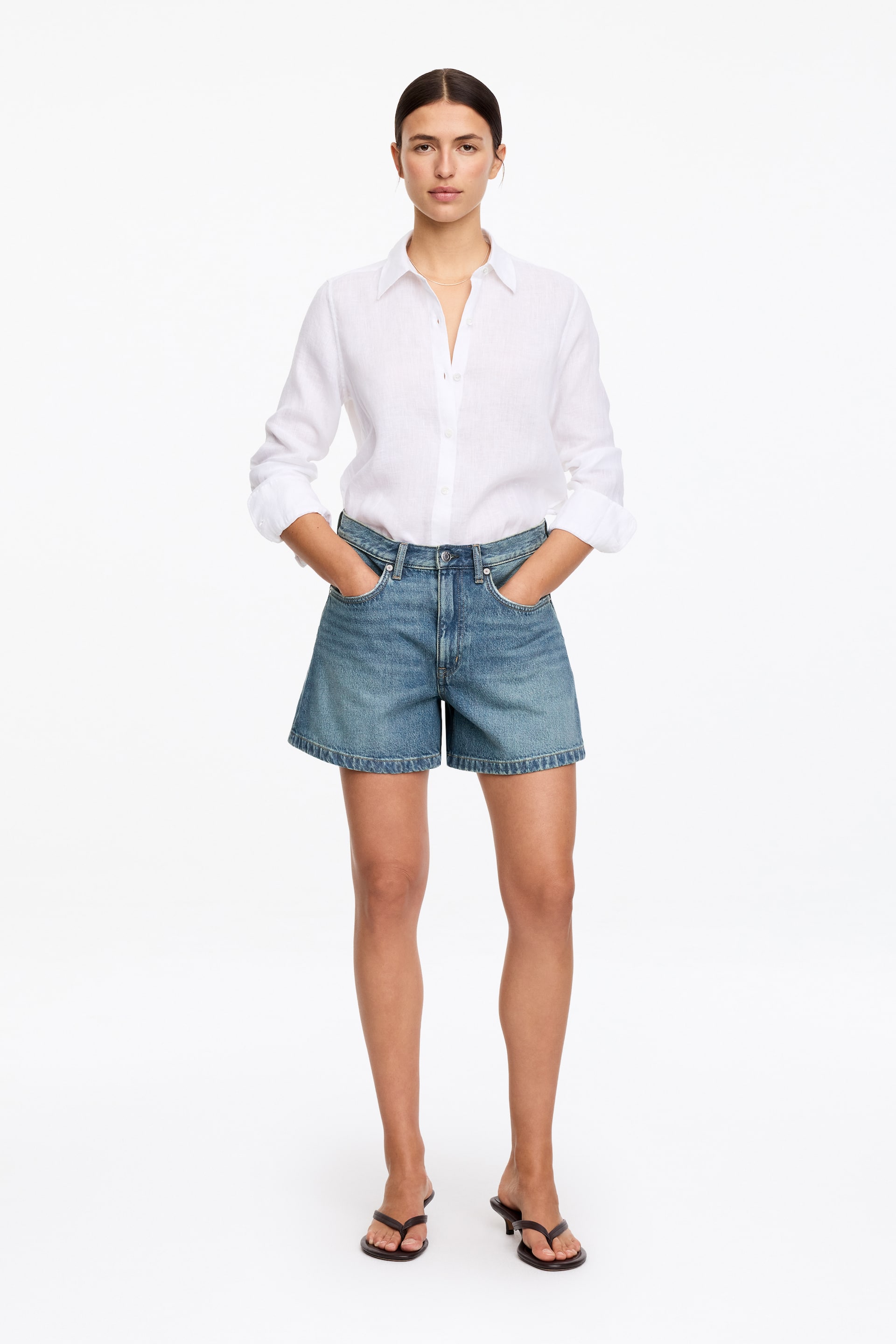 Agrandir l'image: Une chemise blanche en lin à boutons avec des manches longues retroussées et un col classique est rentrée dans un short en jean bleu moyen, qui présente une coupe ample et décontractée avec des poches avant visibles. Des sandales sombres à lanières avec un talon bas complètent l'ensemble.