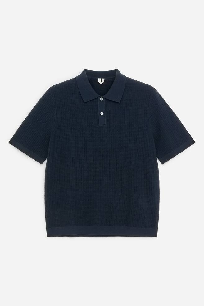 A dark blue knitted polo shirt is showcasing its front, featuring a structured moss stitch throughout the body, short sleeves, and a classic polo collar. It is detailed with a two-button half placket, displaying light-coloured, round buttons, and finished with plain knit bands at the hem and sleeve cuffs. - 1