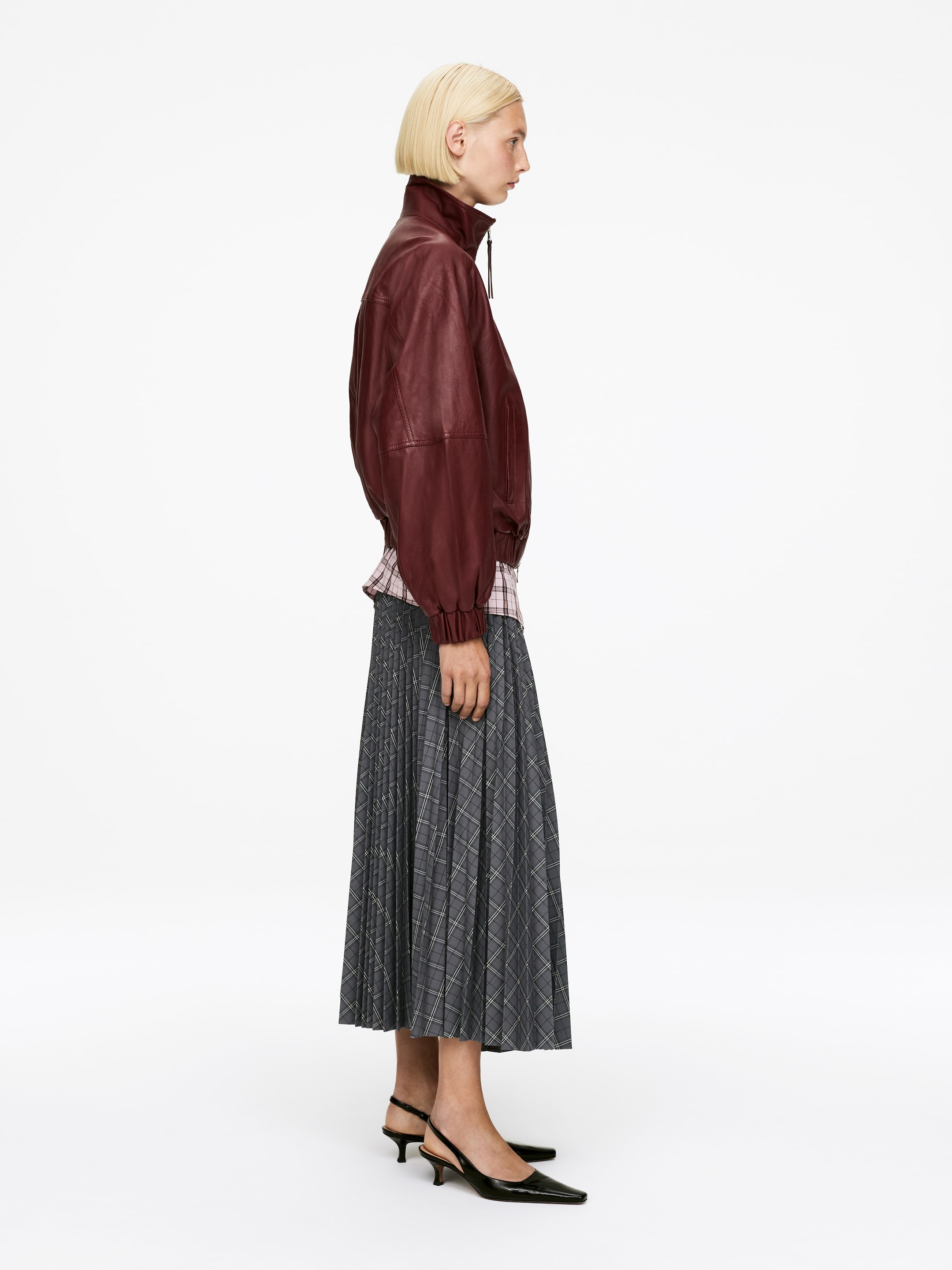 Größeres Bild ansehen: Eine dunkelrote Lederblousonjacke mit Stehkragen, gerafften Bündchen und elastischem Saum wird in einer Seitenansicht über einem hellrosa karierten Hemd präsentiert. Ein grauer, plissierter Midirock mit Karomuster wird ebenfalls getragen, zusammen mit schwarzen Slingback-Kitten-Heels mit spitzer Zehenpartie.