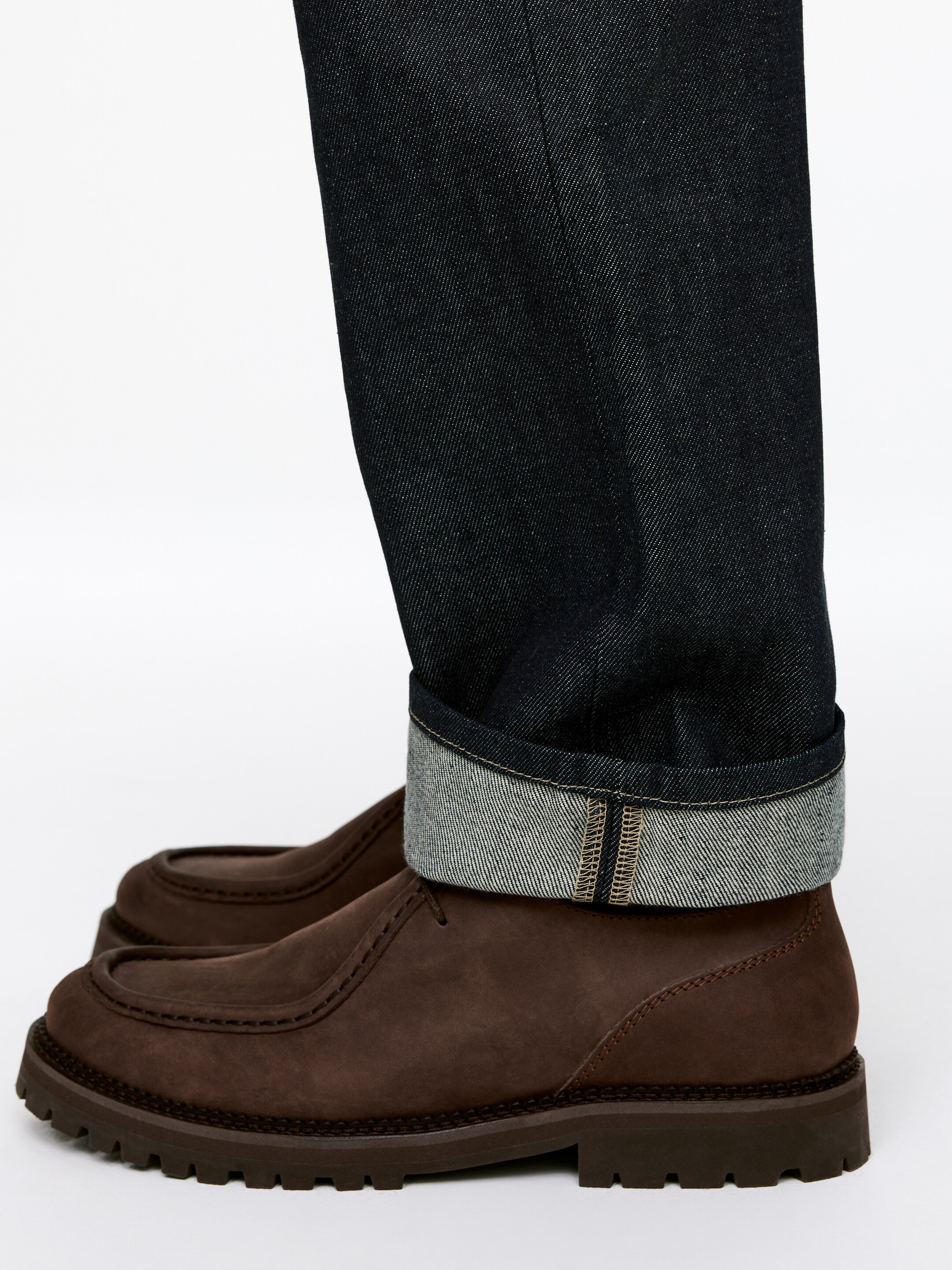 Visualizza immagine più grande: Jeans selvedge indaco scuro sono risvoltati, rivelando un bordo selvedge a linea rossa, e vengono indossati con scarpe moc-toe in camoscio marrone con una spessa suola a carrarmato. Le scarpe sono leggermente angolate verso il fronte.