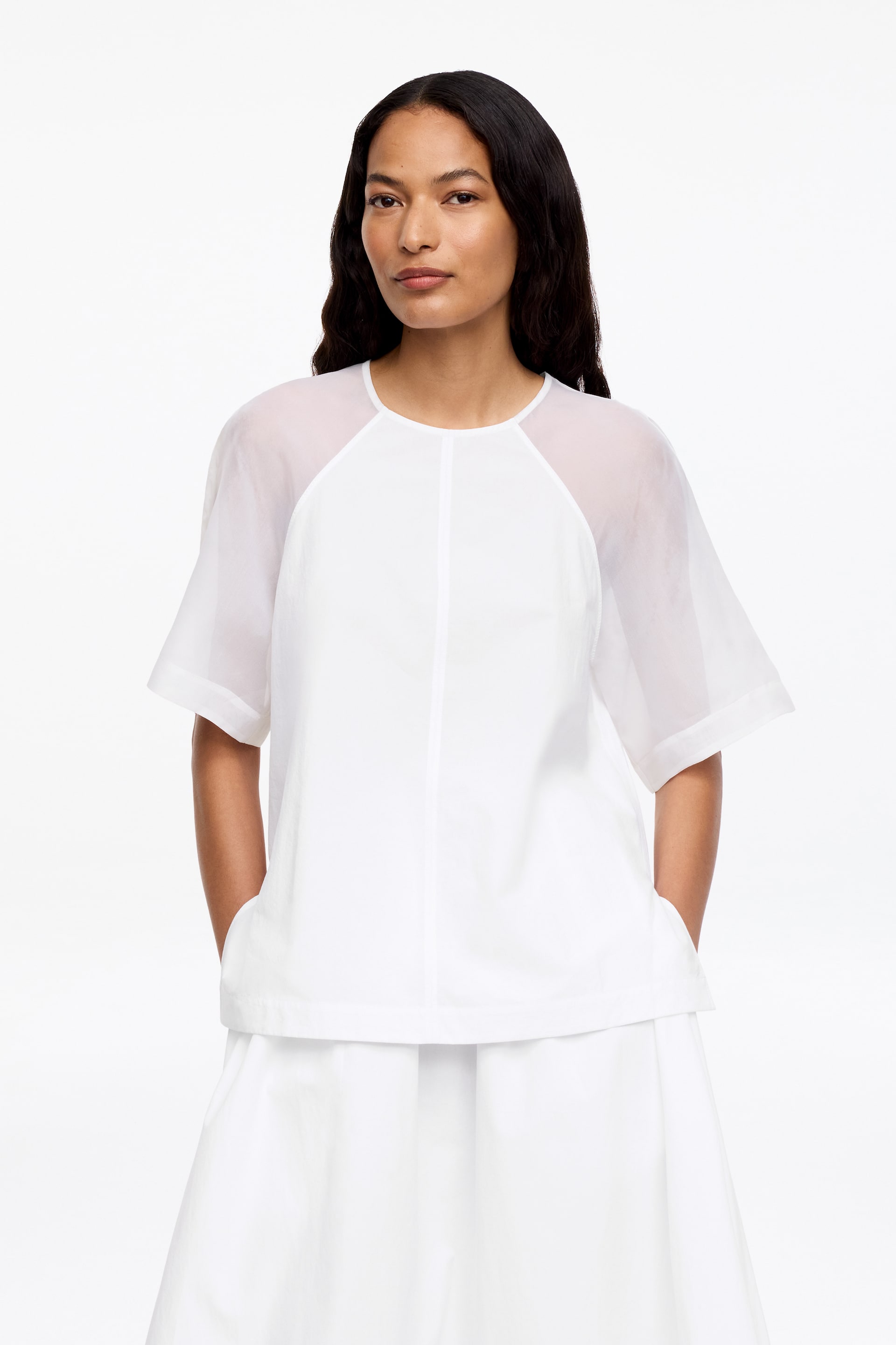 Agrandir l'image: Un haut blanc à manches courtes est porté, avec un col rond, des manches raglan transparentes et un corps opaque avec une couture centrale verticale à l'avant. L'ourlet droit du haut tombe sur une jupe blanche.