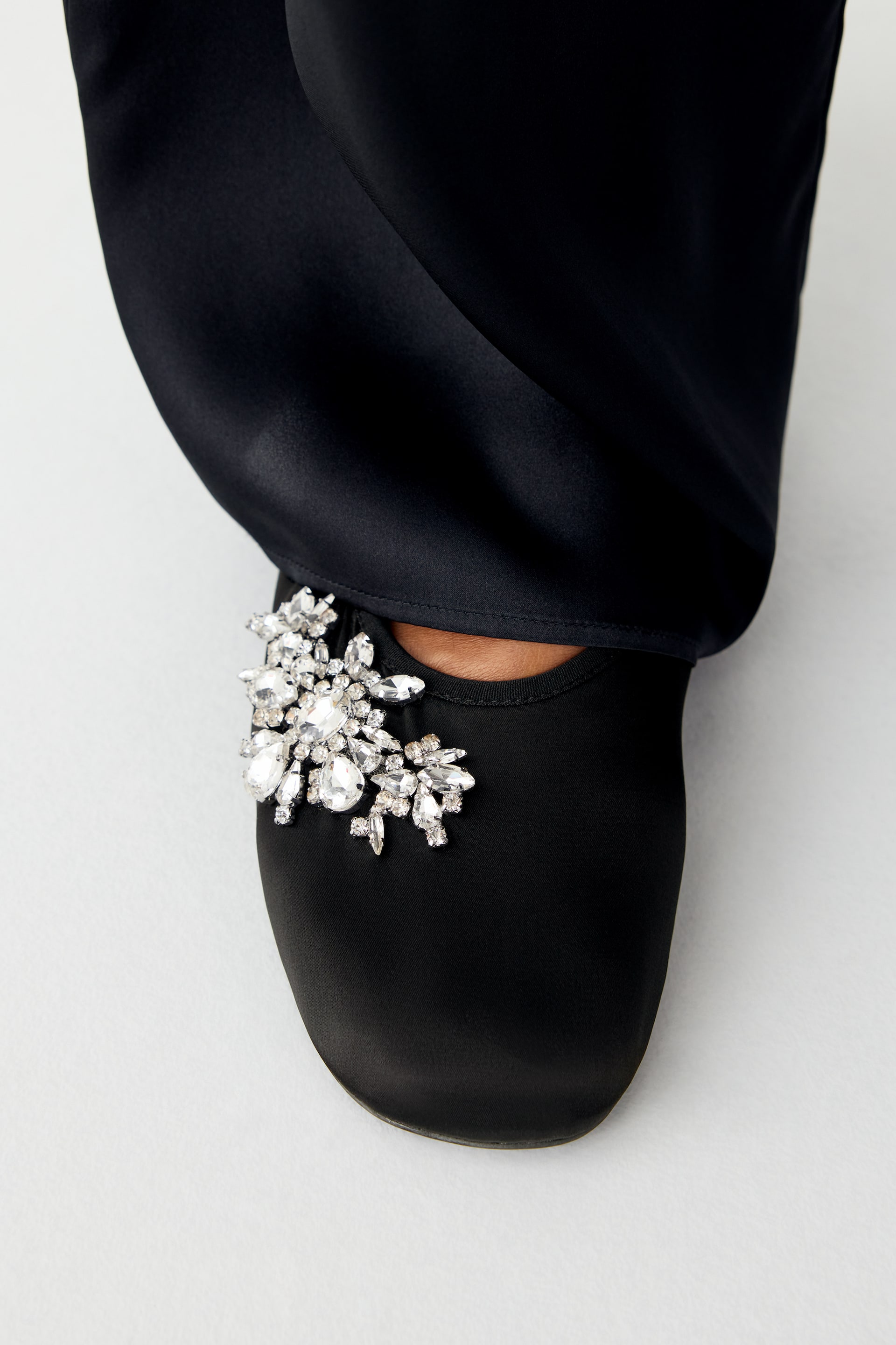 View larger image: The model is wearing a black stretch-satin ballet flat with a gathered edge and a decorative cluster of sparkling glass beads at the toe. The shoe is styled with wide-leg black satin trousers that drape over the ankle.