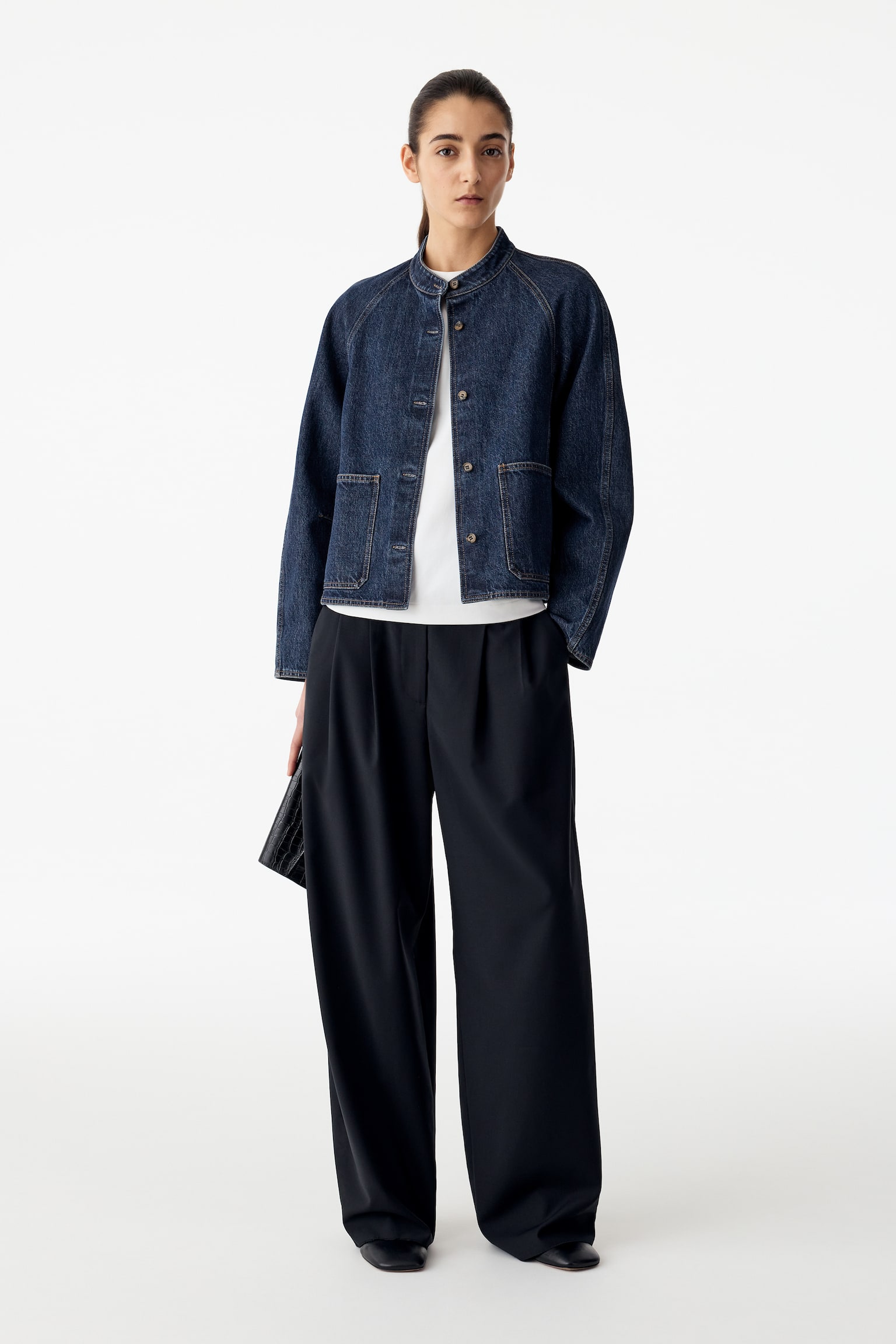 La modella indossa un sovracamicia in denim blu scuro con colletto alla coreana, tasche applicate frontali e chiusura con bottoni. La sovracamicia è indossata sopra una T-shirt bianca ed è abbinata a pantaloni scuri a gamba larga. - 1