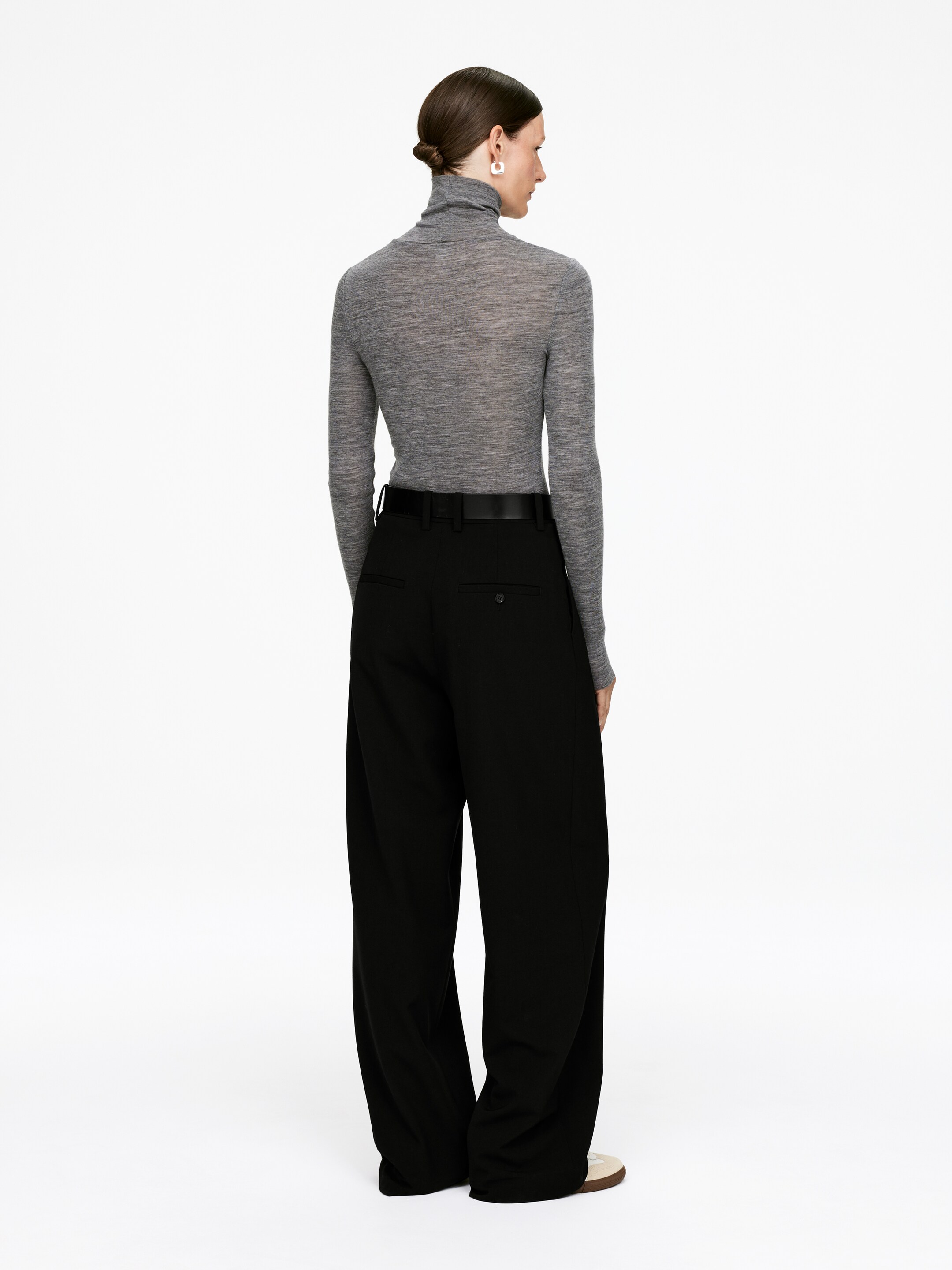 Vis større bilde: En person har på seg en tettsittende, lett grå turtleneck og løse svarte bukser med vide ben, sett bakfra. Turtlenecken sitter tett, mens buksene har en avslappet, flytende drapering med et svart belte rundt midjen.