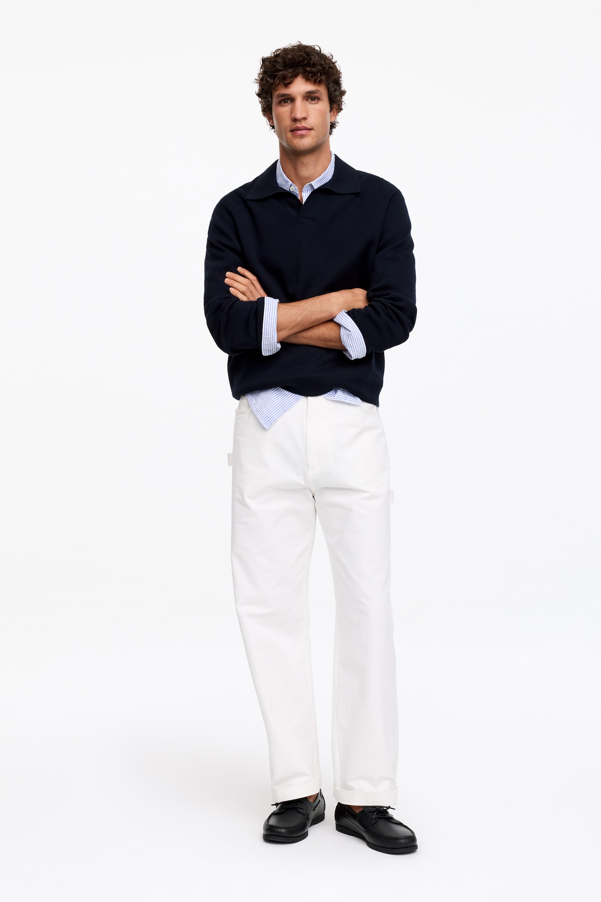 Nagyobb kép megtekintése: Egy sötétkék hosszú ujjú póló pulóver rétegezve van egy világoskék és fehér csíkos galléros ing fölé. Fehér, egyenes szárú nadrággal és fekete fűzős hajócipővel van kombinálva, teljes elölnézetből bemutatva.