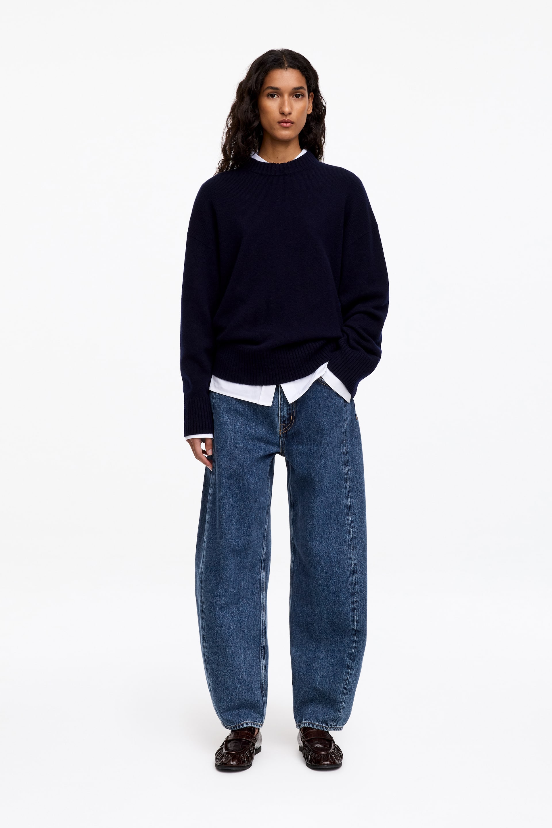 Visualizza l'immagine ingrandita: Jeans in denim blu con vestibilità rilassata e gamba scolpita e curva, indossati dalla modella. I jeans sono abbinati a un maglione girocollo navy portato sopra una camicia bianca con colletto e mocassini marrone scuro.