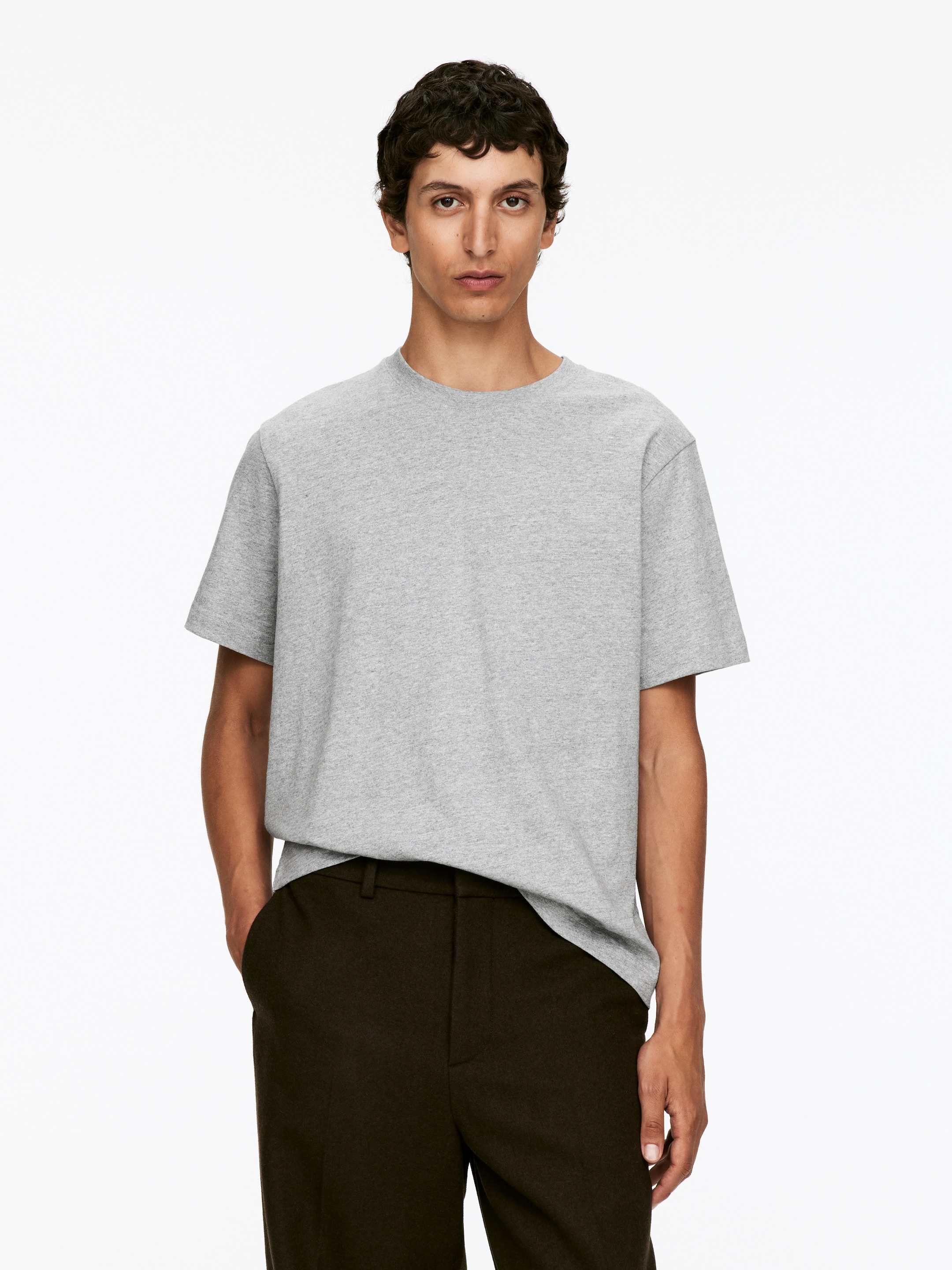 Visualizza immagine più grande: Una t-shirt a maniche corte grigio chiaro è presentata su una persona rivolta in avanti. Presenta un classico scollo a girocollo e una vestibilità rilassata, con il tessuto che drappeggia delicatamente e viene indossato fuori dai pantaloni scuri.
