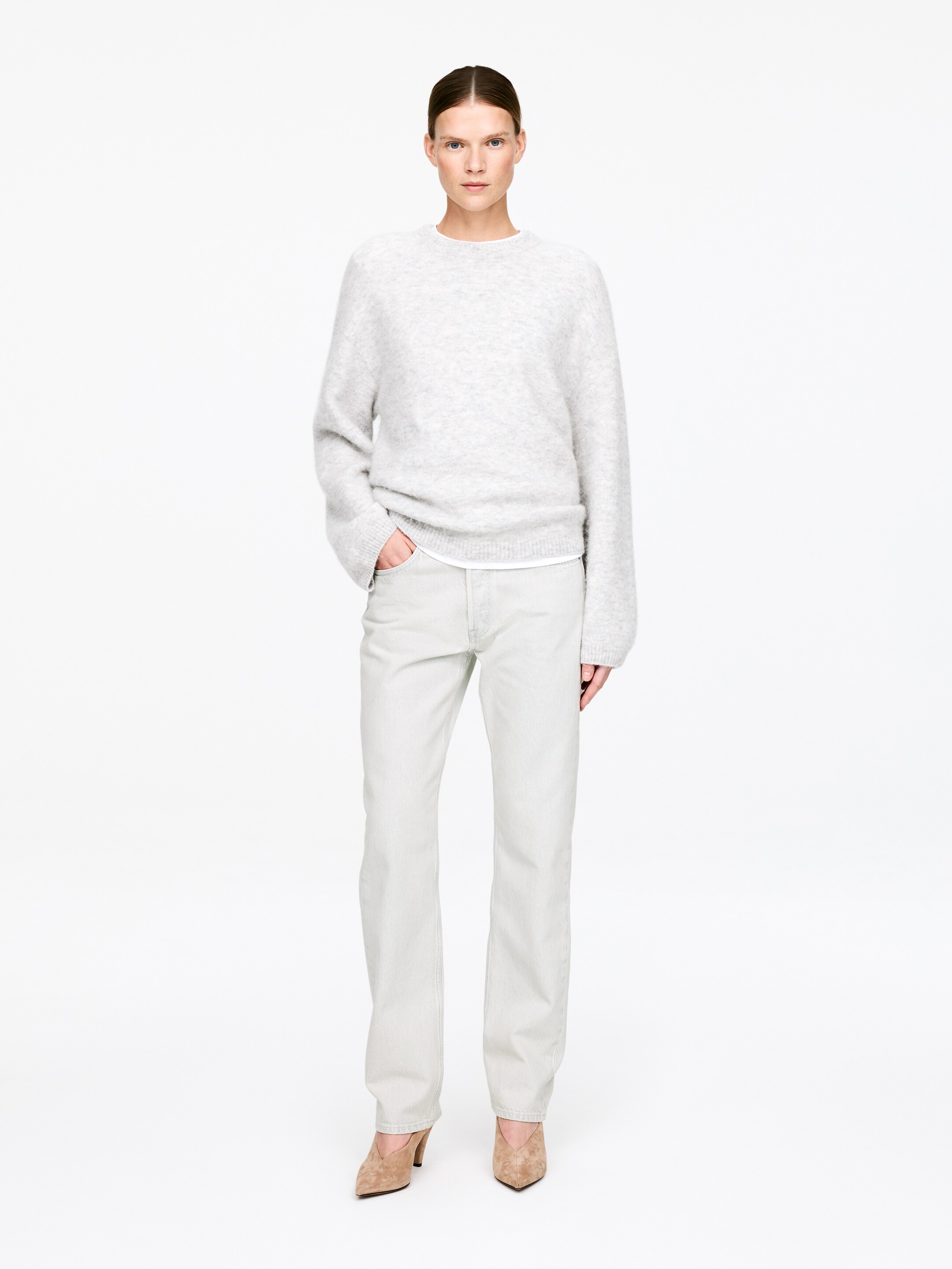 Προβολή μεγαλύτερης εικόνας: Ένα ανοιχτό γκρι πλεκτό πουλόβερ με στρογγυλή λαιμόκοψη προσφέρει μια χαλαρή εφαρμογή, διαθέτοντας μακριά μανίκια και ριγέ τελείωμα. Ένα λευκό t-shirt είναι ορατό σε στρώσεις από κάτω, εμφανιζόμενο στη λαιμόκοψη και στο κάτω τελείωμα.