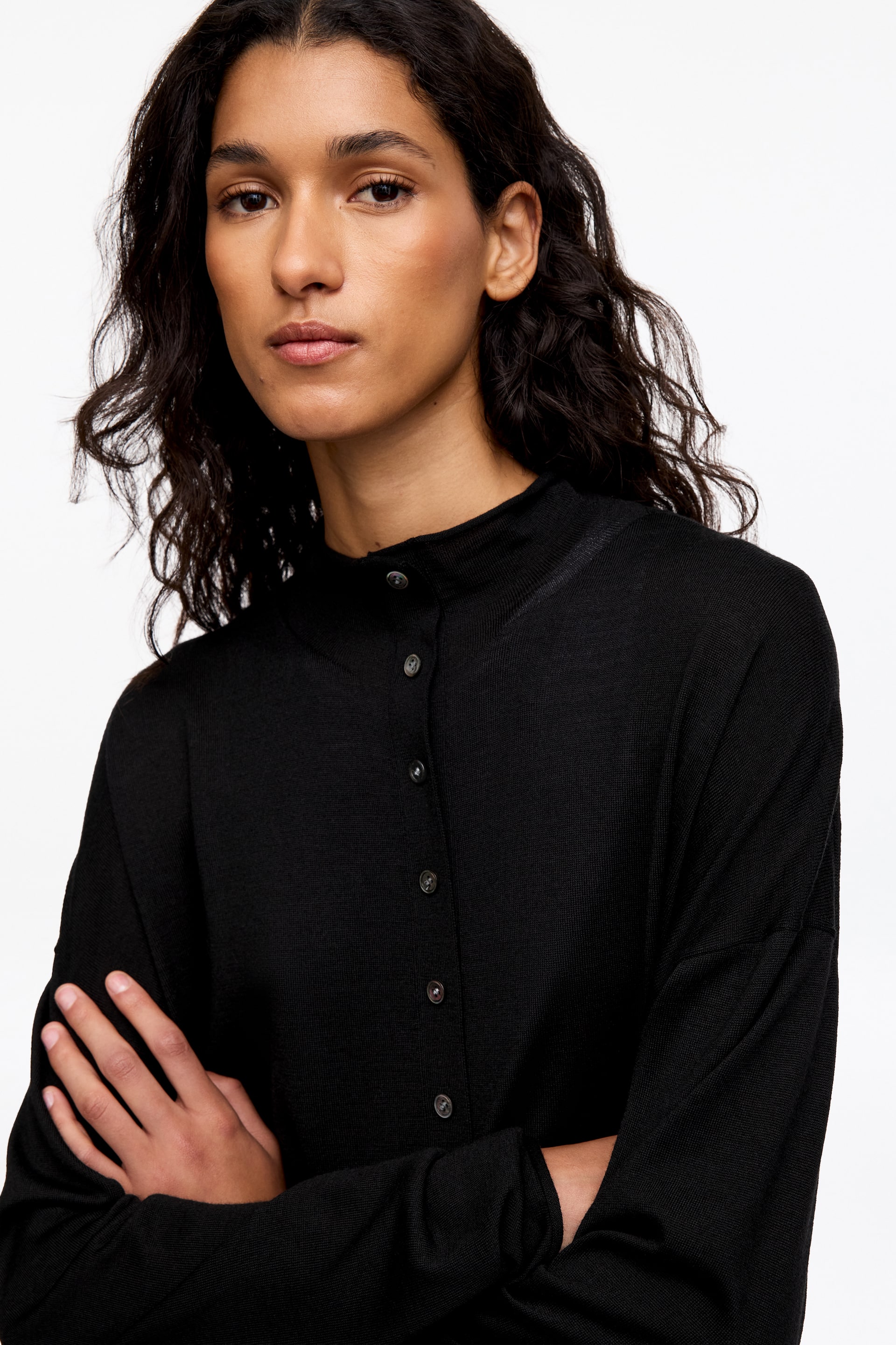 Agrandir l'image: Le mannequin porte un cardigan noir en mélange de laine avec col montant, en prenant la pose les bras croisés. Le cardigan présente une fermeture boutonnée sur le devant et un col montant haut de type mock-neck.