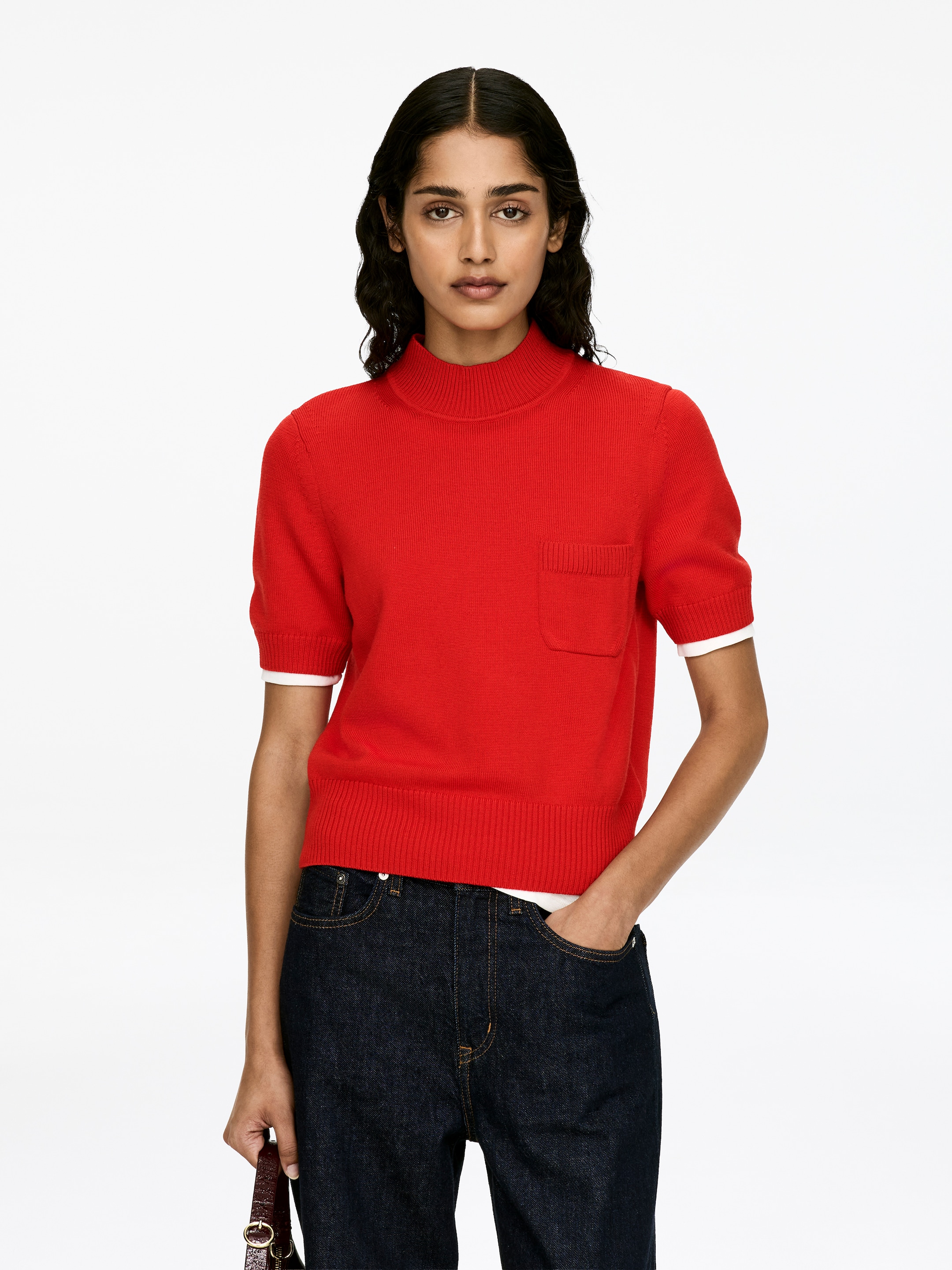 Visa större bild: En röd, kortärmad tröja med en mock-neck, en bröstficka och ribbstickade muddar och fåll, bärs av modellen över ett synligt vitt lager. Modellen bär också mörka jeans.