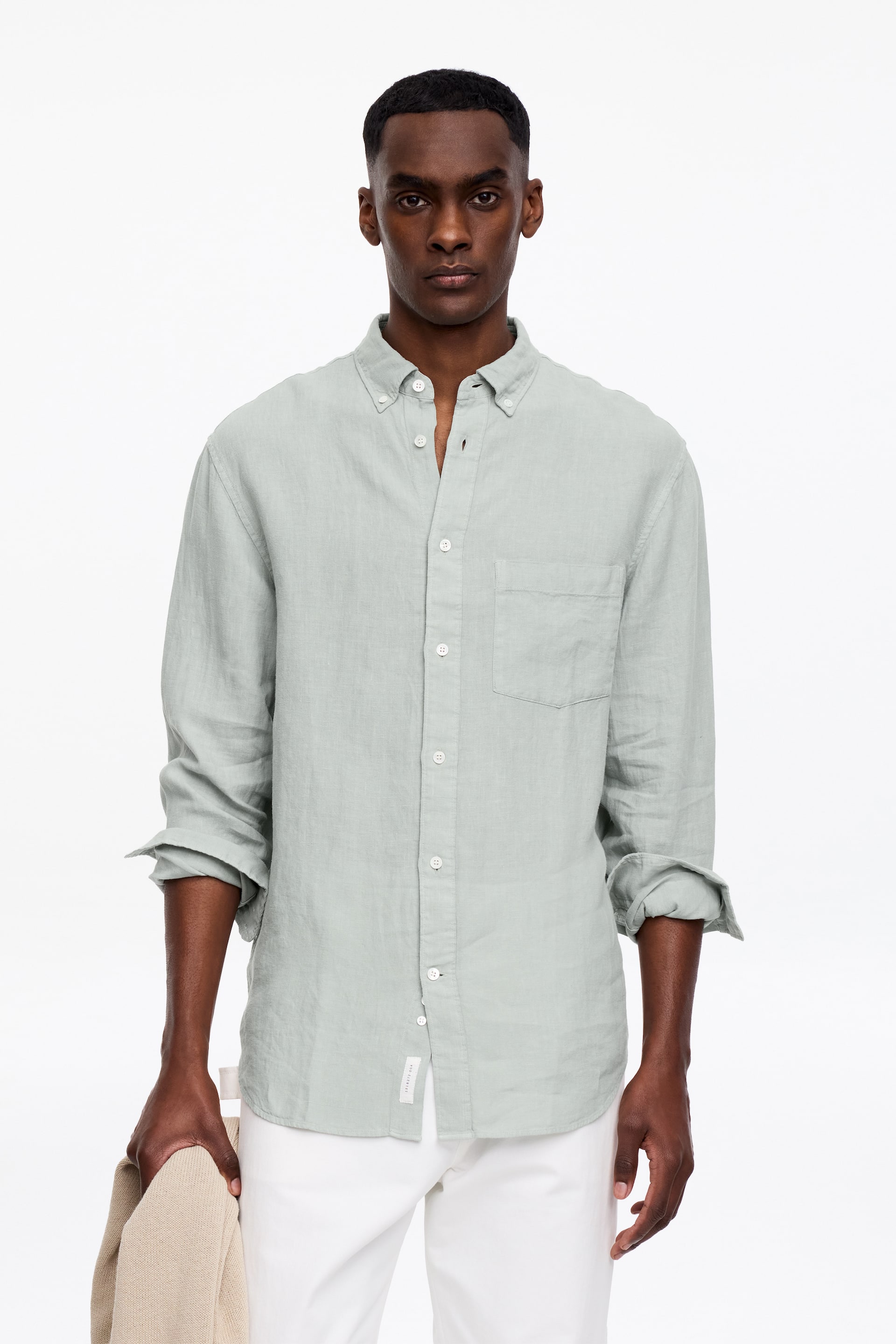 Zobacz większy obraz: Jasnozielona lniana koszula jest prezentowana przodem, z kołnierzykiem zapinanym na guziki i pełnym rzędem guzików z przodu z białymi guzikami. Posiada pojedynczą kieszeń naszywaną na lewej piersi, a jej długie rękawy są podwinięte do przedramion.