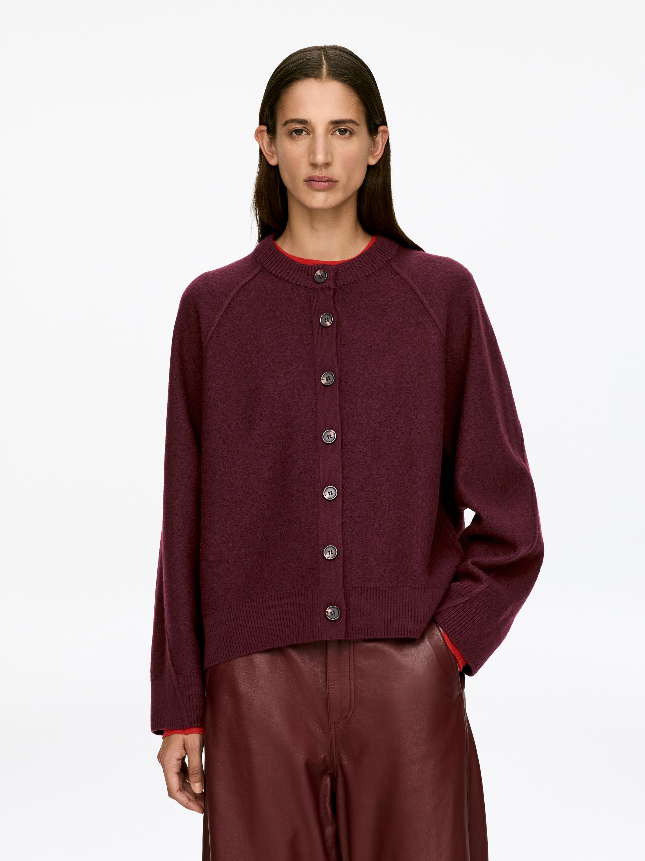 Agrandir l'image: Un cardigan en laine bouillie rouge foncé, avec un col rond, des manches raglan, une fermeture à boutons avec cinq boutons foncés, et des bordures côtelées au col, aux poignets et à l'ourlet, est porté par le mannequin. Le cardigan ample est superposé sur un haut rouge, visible au niveau de l'encolure et des poignets.