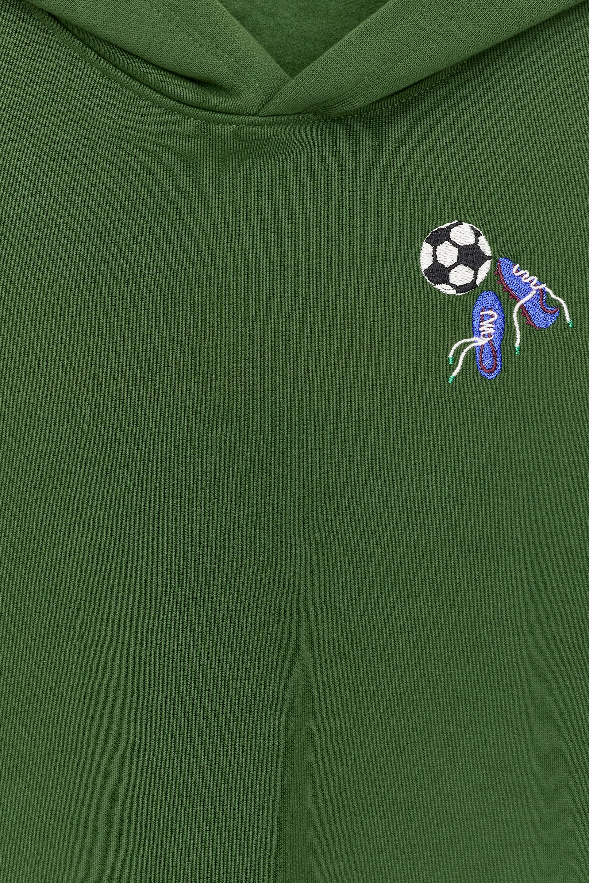 Ver imagen más grande: Una sudadera con capucha verde muestra un estampado bordado en el pecho, con un balón de fútbol en blanco y negro y dos botas de fútbol azules con cordones blancos.