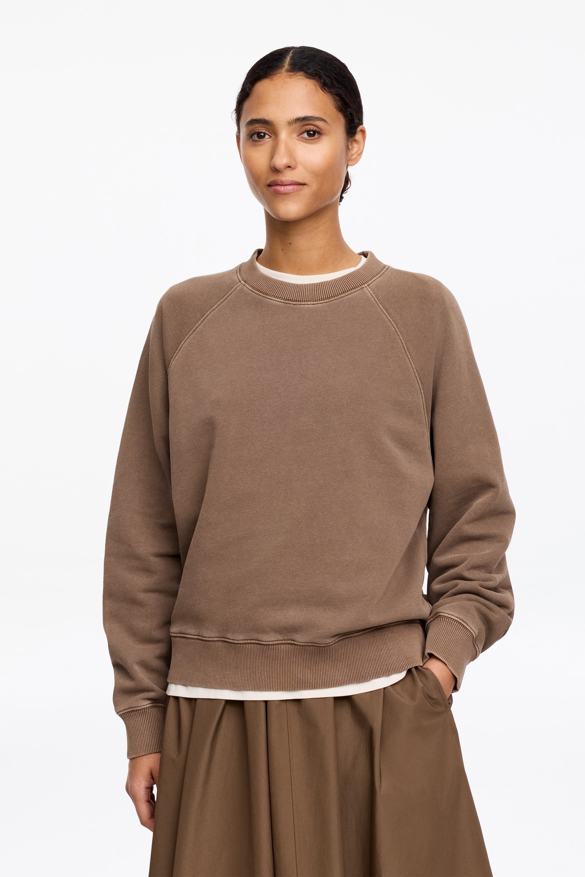 Ingrandisci l'immagine: Una felpa marrone con scollo a girocollo è presentata, caratterizzata da maniche raglan con cuciture visibili, polsini a coste e un orlo a coste. Una t-shirt bianca con scollo a girocollo è stratificata sotto, estendendosi oltre l'orlo della felpa.