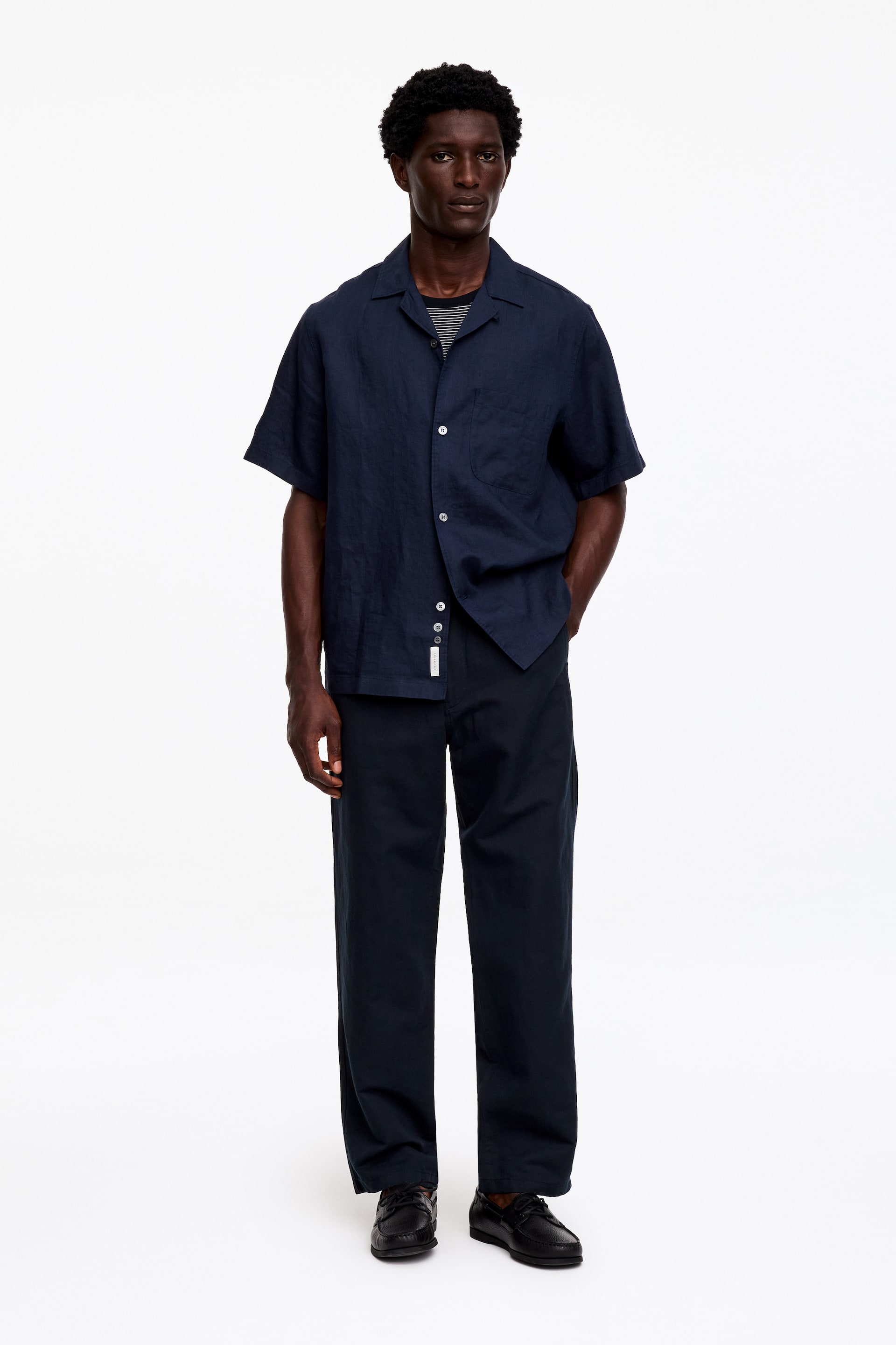 Größeres Bild ansehen: Ein Outfit mit einem marineblauen Kurzarmhemd mit Camp-Kragen, das offen über einem schwarz-weiß gestreiften T-Shirt getragen wird, dazu passende marineblaue gerade Hosen am Unterkörper und schwarze Leder-Loafer, die das Ensemble vervollständigen. Das gesamte Outfit wird frontal präsentiert.