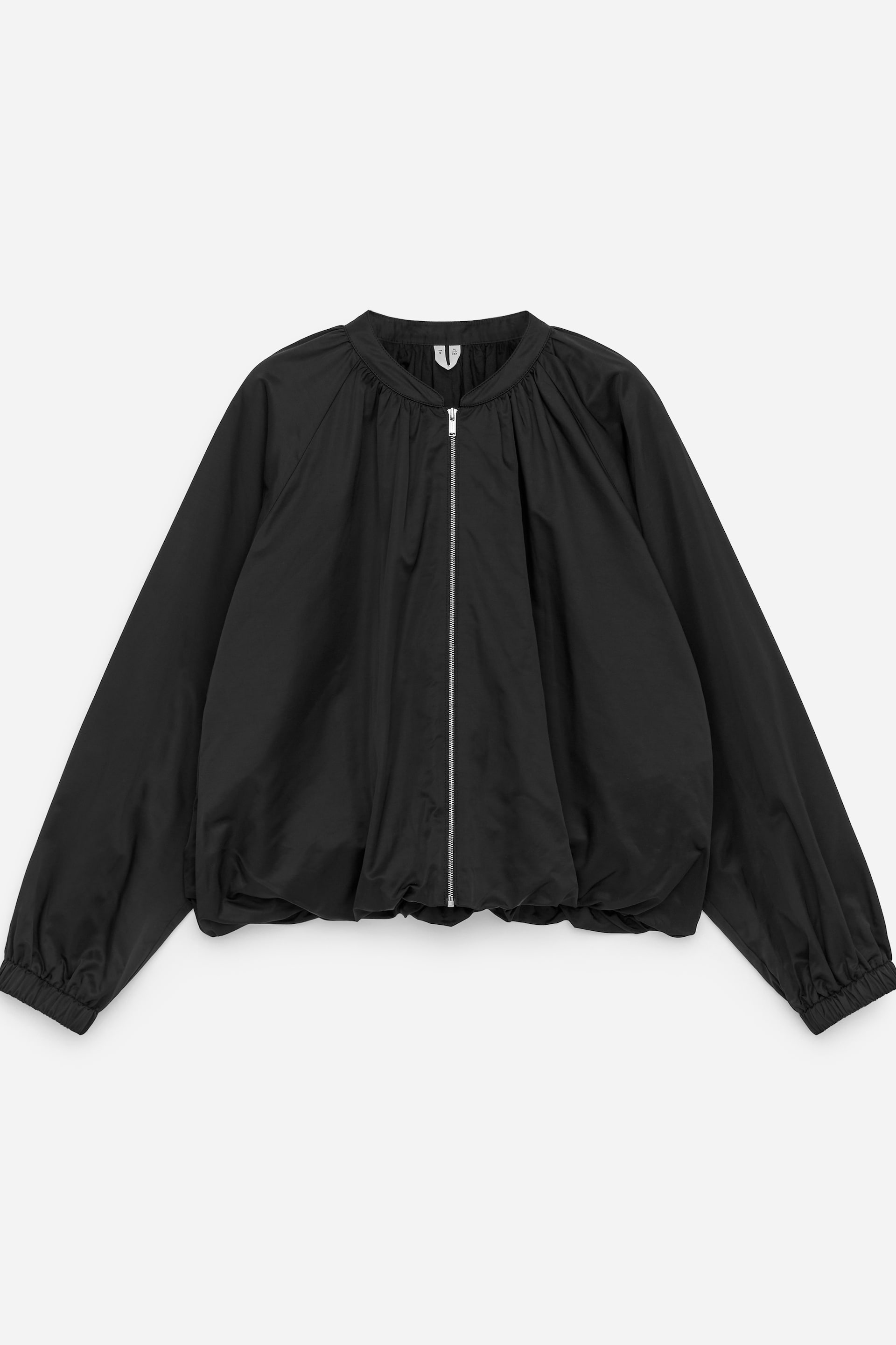 Größeres Bild ansehen: Eine schwarze Jacke wird von vorne präsentiert, hergestellt aus einem glatten Satin-Nylon-Material. Sie verfügt über einen Stehkragen, einen durchgehenden Reißverschluss, sanfte Raffungen am Hals, die eine subtile ballonartige Form erzeugen, sowie elastische Bündchen und einen elastischen Saum.