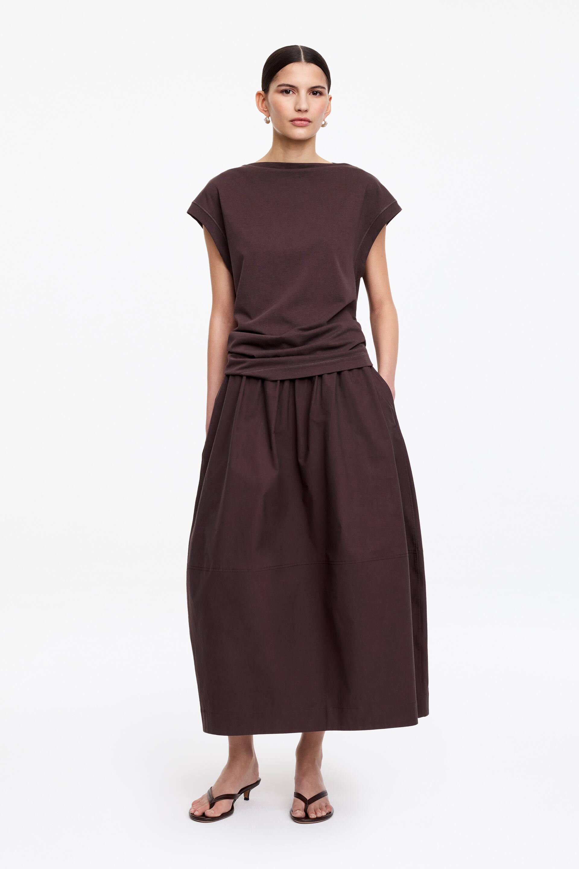 Nagyobb kép megtekintése: Egy sötétbarna, csónaknyakú, rövid ujjú felsőt viselnek, amelynek szegélye egy hozzáillő sötétbarna, oldalzsebes és vízszintes varrással ellátott, rakott midi szoknyába van tűrve.