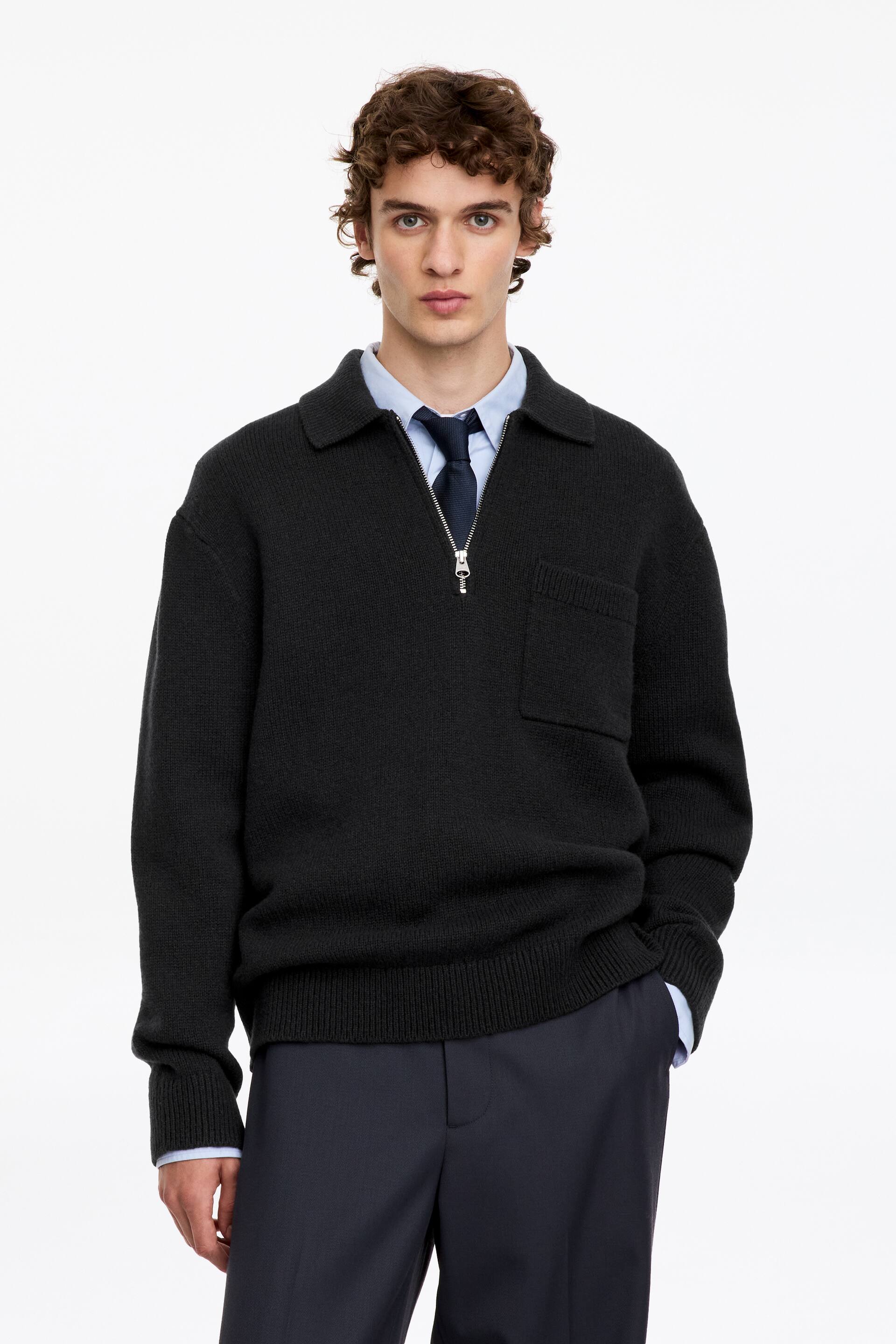 View larger image: A black half-zip knitted sweater featuring a collar and a single chest pocket is worn by the model. The sweater is layered over a light blue collared shirt with a dark tie and paired with dark trousers.
