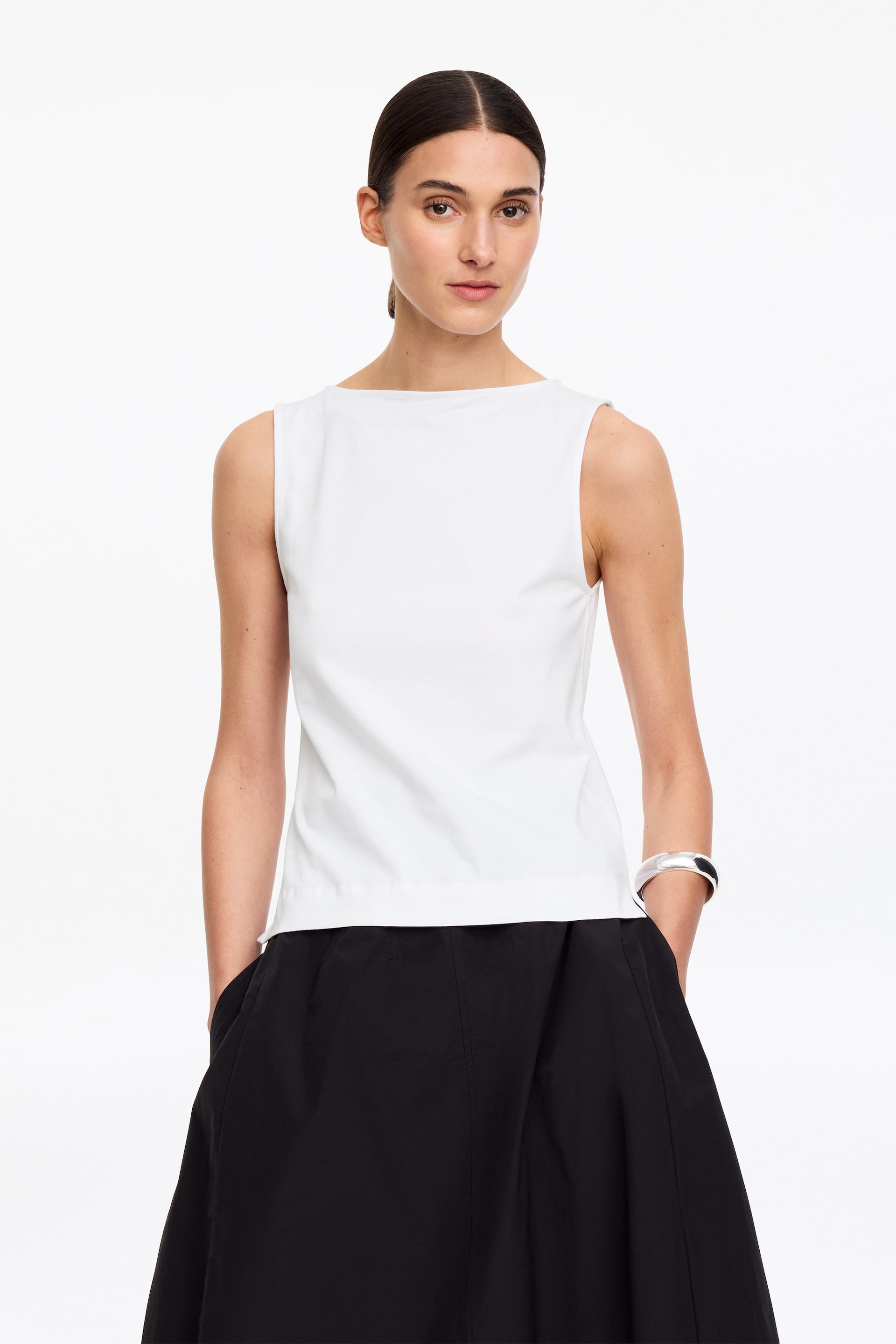 Ingrandisci l'immagine: Un top bianco senza maniche presenta uno scollo a barca e un orlo dritto. Offre una vestibilità rilassata e una texture liscia, mentre si mostra frontalmente.