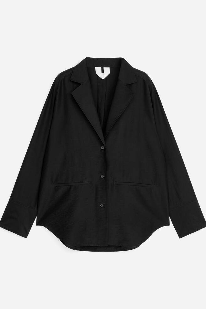 En svart blazer vises fra forsiden, med spisse slag og en knappet frontlukking. Den har lange ermer med brede mansjetter, to rektangulære lommeflapper og en avrundet kant. - 1