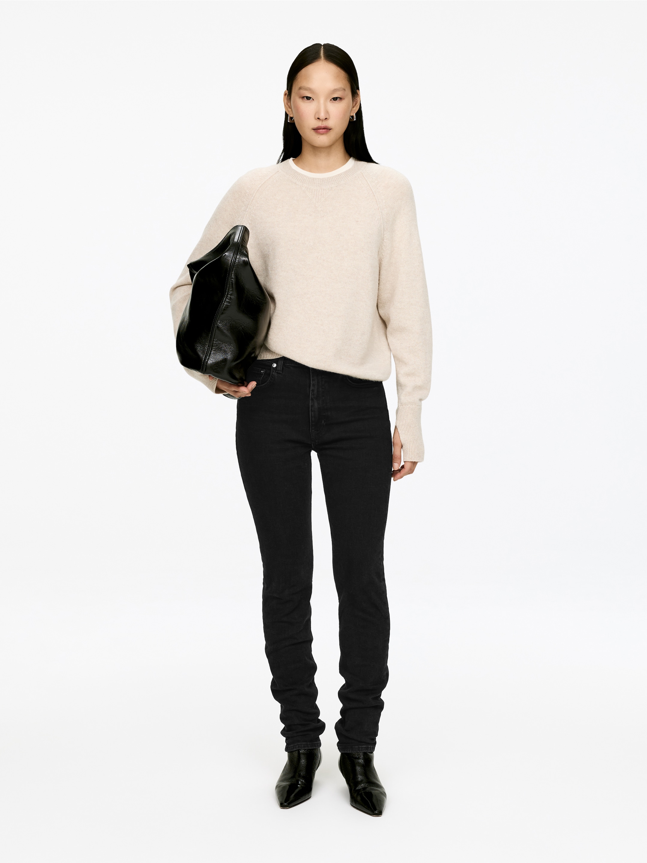 Visa större bild: En ljusbeige tröja med rund hals och raglanärmar samt ribbade muddar, är stylad front mot front med svarta skinny jeans. En vit t-shirt syns från tröjans halslinje, medan en svart slapp väska hålls, och svarta ankelboots med spetsiga tår bärs.