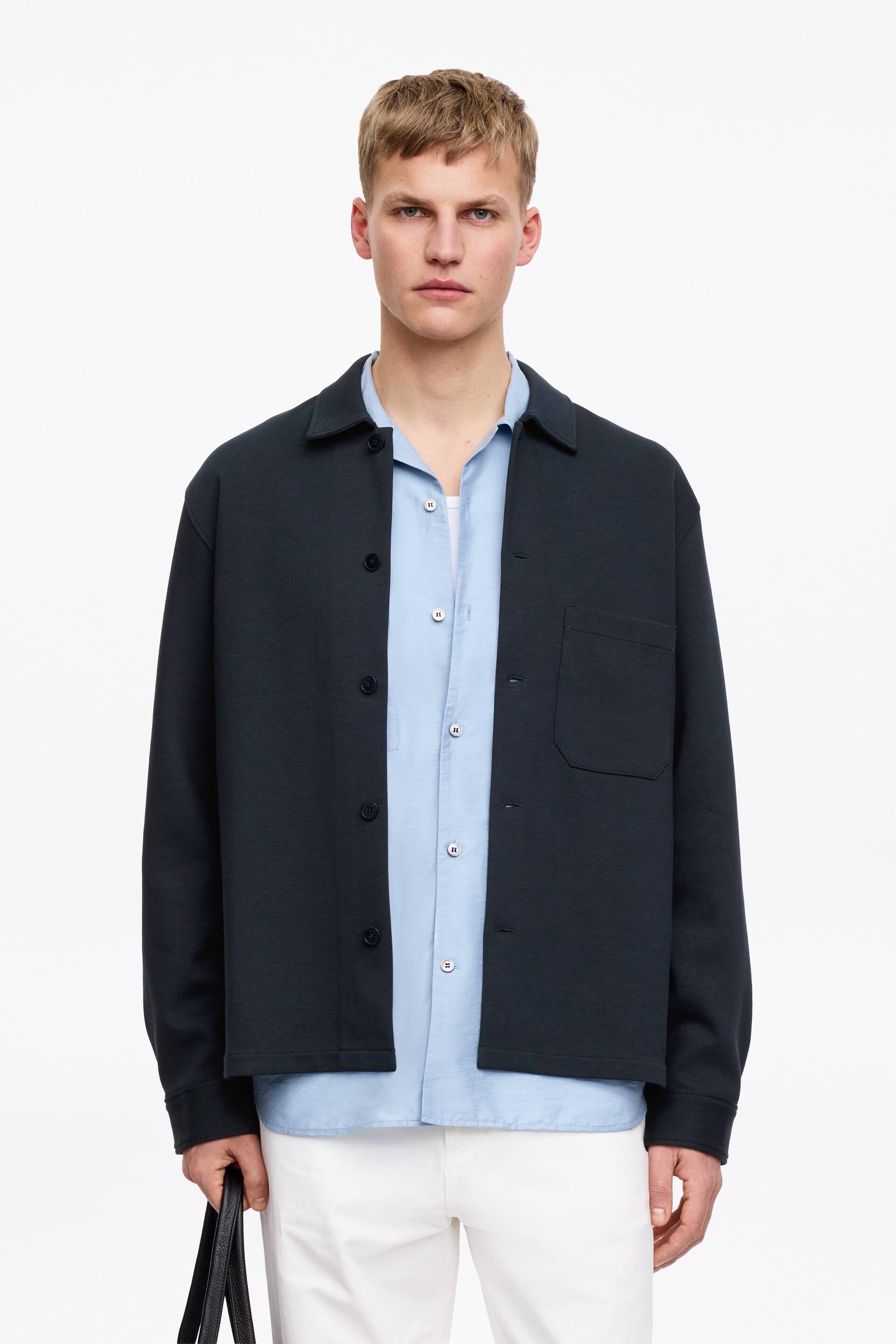 Преглед на по-голямо изображение: Тъмносин памучен джърси оувършърт с нормална кройка, със стандартна яка, предно закопчаване с копчета и един джоб на гърдите, моделът го носи разкопчан върху светлосиня риза с копчета.