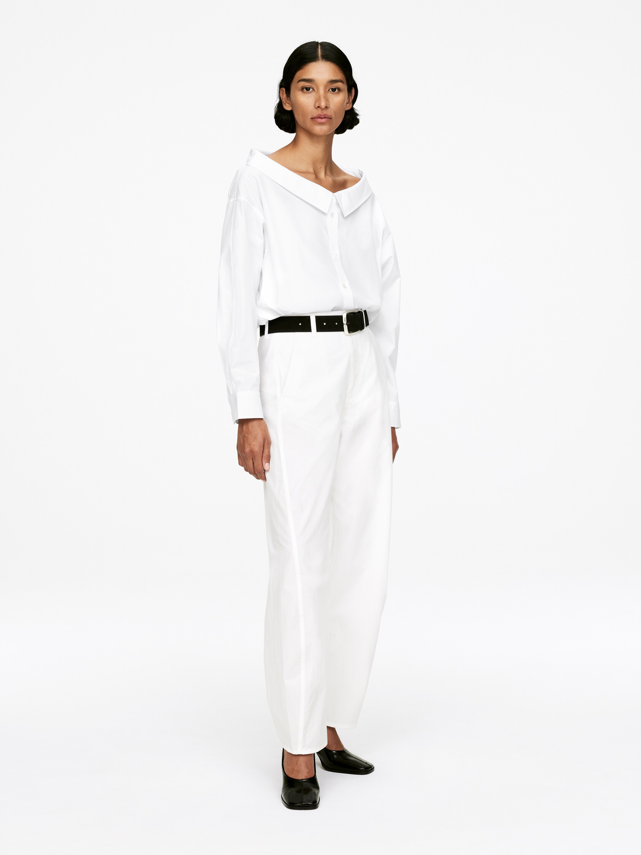 Ingrandisci l'immagine: Una camicia bianca con bottoni è indossata, caratterizzata da uno scollo aperto e asimmetrico, stile off-the-shoulder e maniche lunghe. La camicia è infilata in pantaloni bianchi a gamba larga e vestibilità ampia. Una cintura nera stringe la vita sopra i pantaloni, e vengono indossate scarpe piatte nere a punta quadrata.