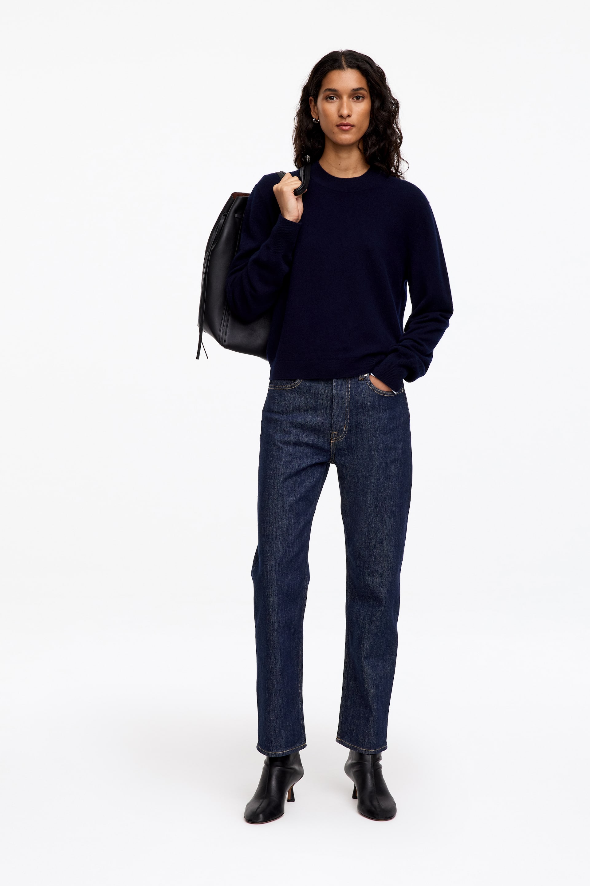 Grösseres Bild ansehen: Dunkelblaue Slim-Fit-Jeans werden vom Model getragen, das mit einer Hand in der vorderen Tasche steht. Die Jeans werden mit einem langärmeligen Marine-Pullover mit Rundhalsausschnitt, schwarzen Stiefeletten mit Absatz und einer schwarzen Lederhandtasche über der Schulter kombiniert.