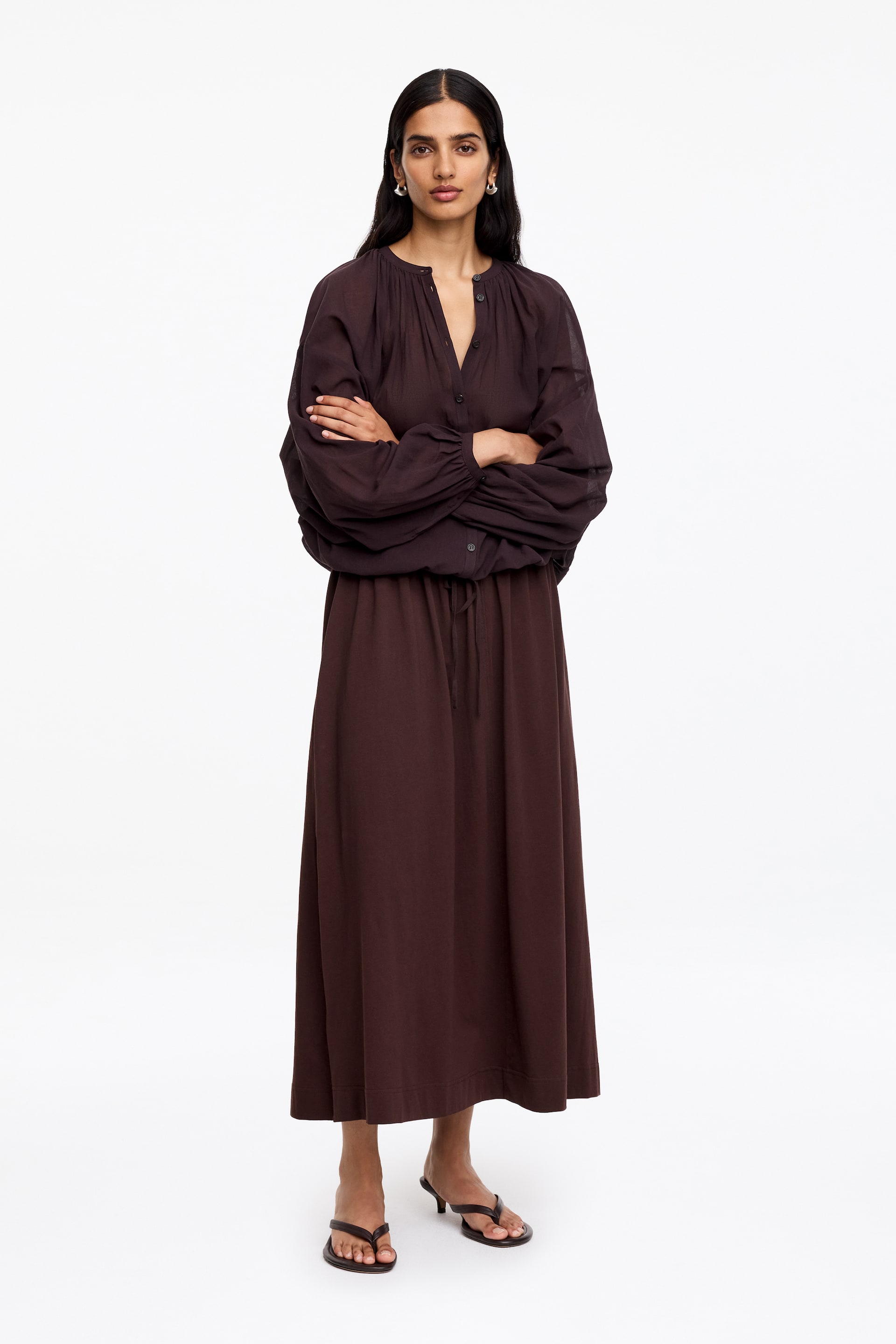 View larger image: A dark brown midi dress featuring a gathered V-neckline with a partial button placket and voluminous sleeves with gathered cuffs. The dress is cinched at the waist with a drawstring, creating a blouson top, and the skirt flows to a midi length.