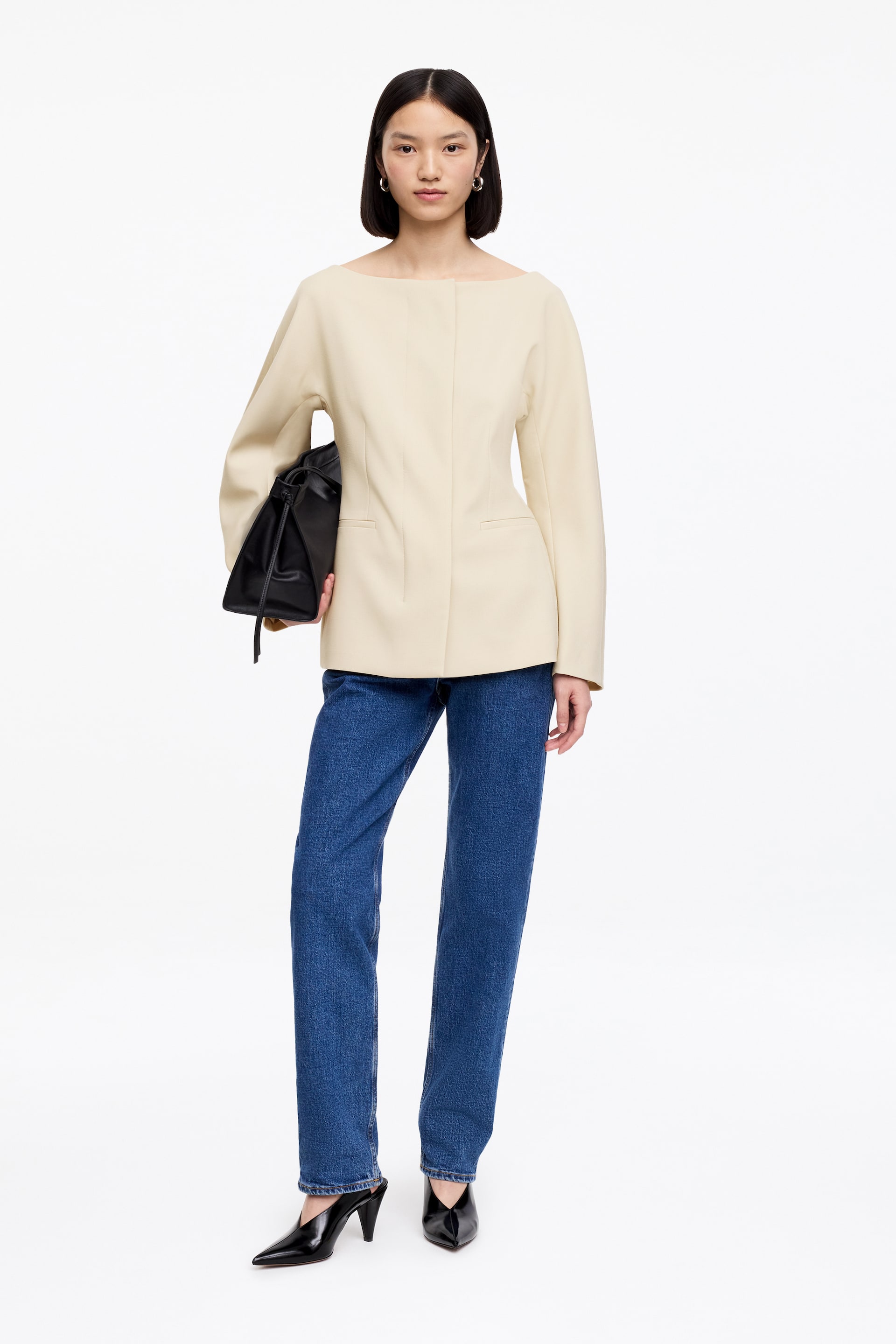 Größeres Bild ansehen: Ein cremefarbenes Oberteil mit einem Bootsausschnitt, langen weiten Ärmeln und einer strukturierten, kastigen Passform. Es verfügt über zwei horizontale Leistentaschen und vertikale Nähte auf der Vorderseite.