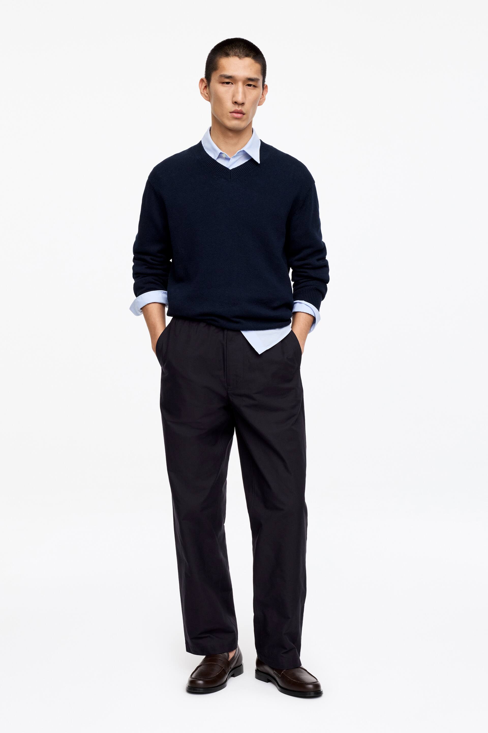 Visualizza l'immagine ingrandita: Un maglione blu navy con scollo a V, caratterizzato da maniche lunghe, polsini a coste e orlo a coste, viene indossato a strati sopra una camicia azzurra con colletto, con il colletto e i polsini visibili.