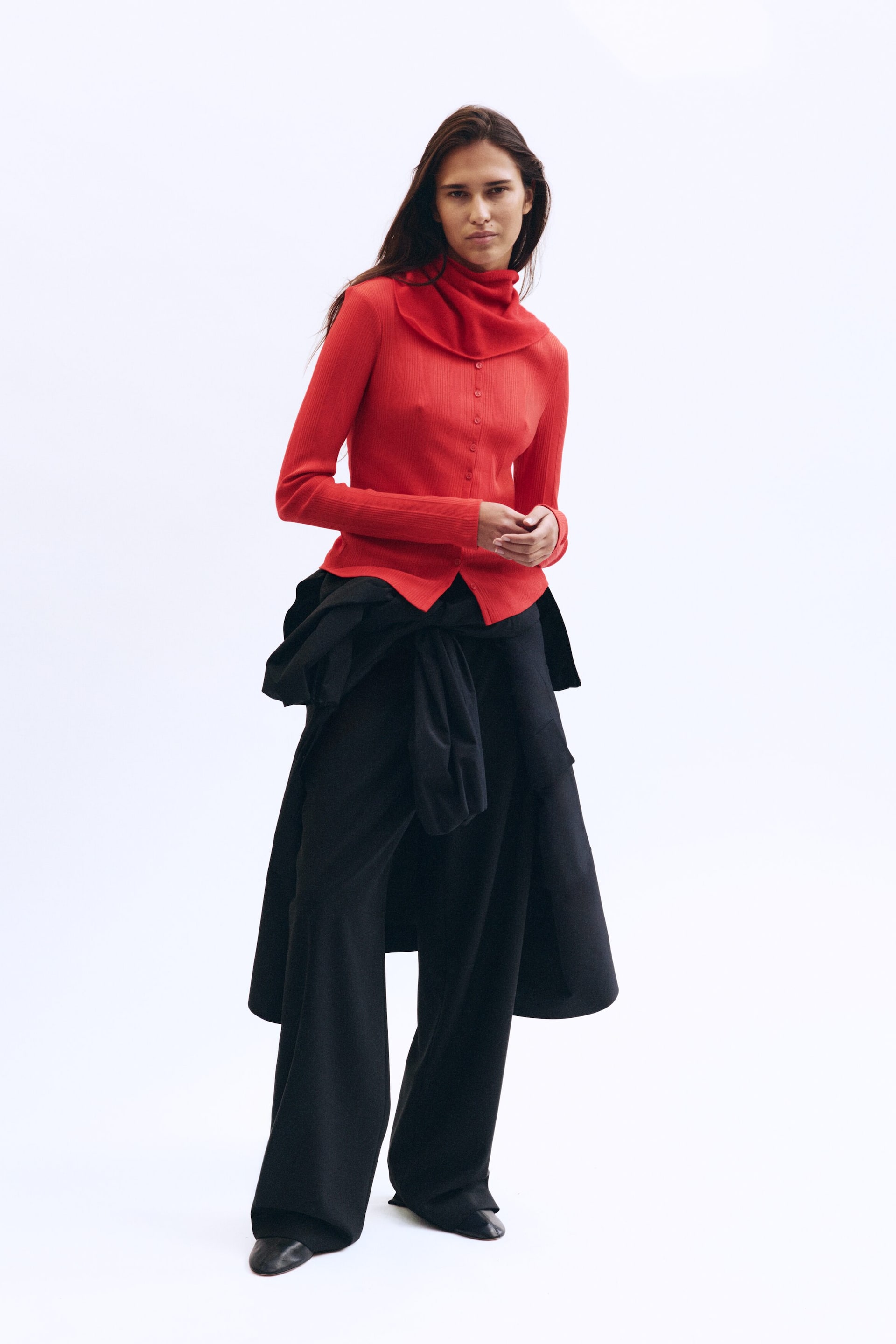 View larger image: Facing front, a model is wearing a bright red ribbed button-front top styled with a matching draped neck collar, paired with relaxed black wide-leg trousers and black leather boots. A black knee-length parka is wrapped and tied around her waist, layered over the trousers.