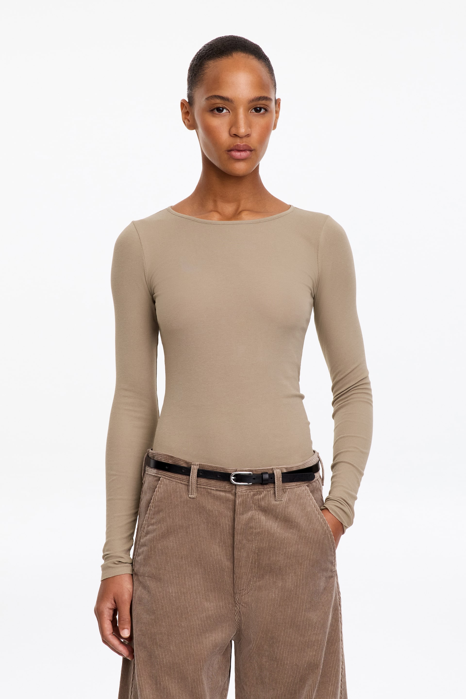 Visualizza l'immagine ingrandita: La modella indossa un top beige, aderente e a maniche lunghe, con scollo rotondo. Il top è infilato in un paio di pantaloni marroni in velluto a coste, abbinati a una sottile cintura nera.