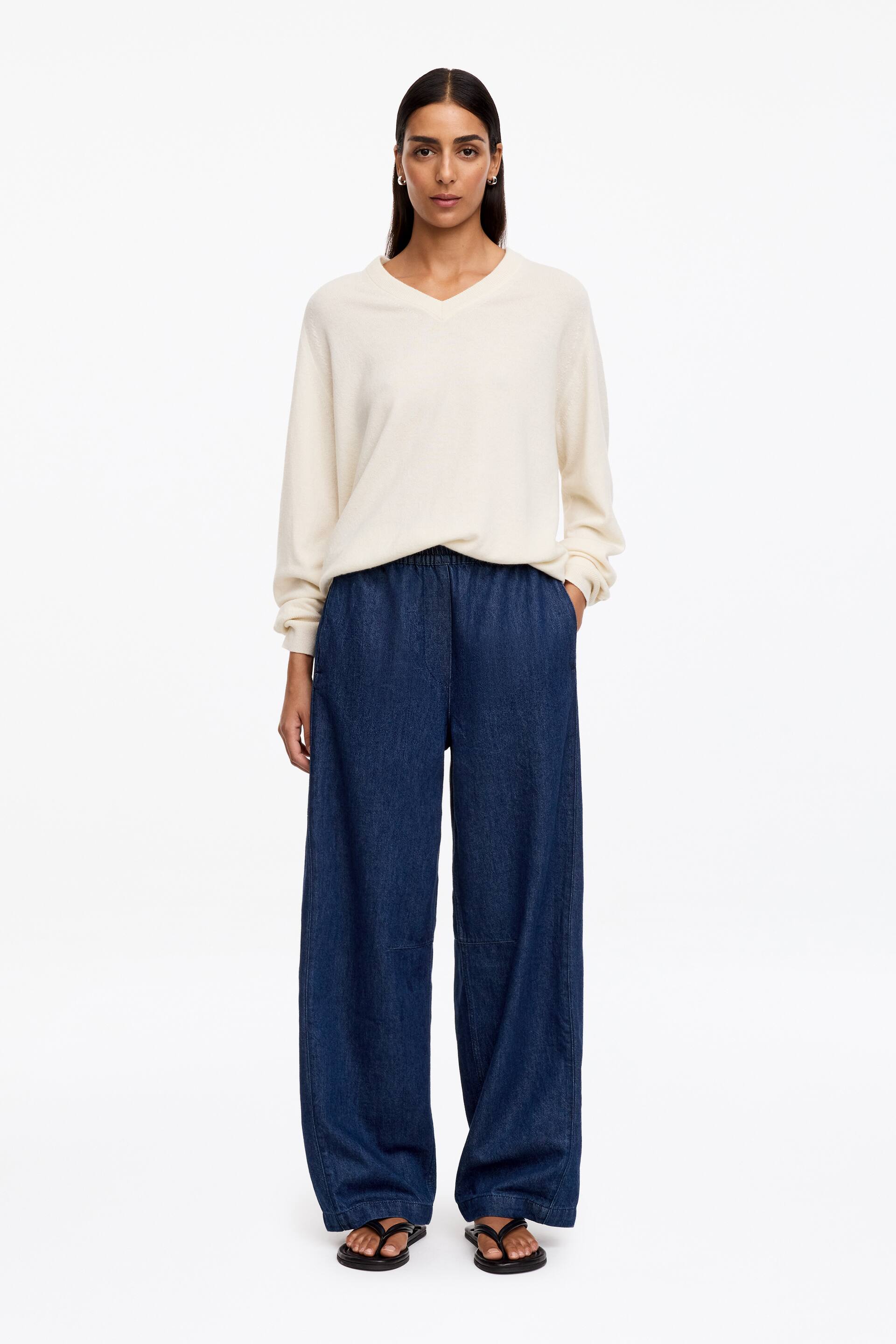 Ver imagem maior: A modelo está a usar umas calças de ganga azul-escuro leves, com uma silhueta relaxed barrel-leg e cintura elástica franzida de vestir, visível por baixo de uma camisola creme. Observam-se detalhes de costura pespontada e um corte subtilmente curvado nas pernas, e a modelo está a colocar uma mão num bolso lateral.